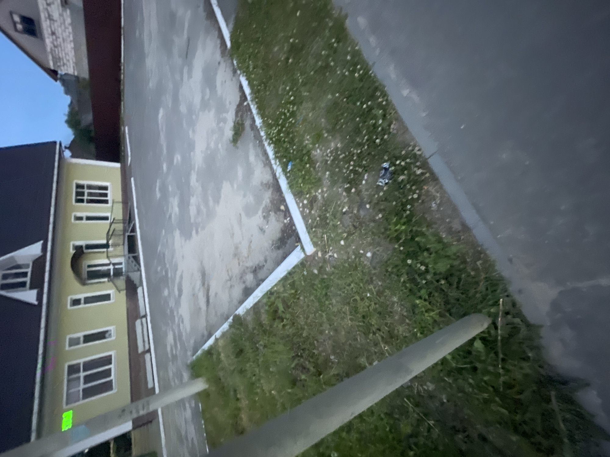 The image depicts a light yellow building with a dark roof, possibly a public facility or residential house, situated in the town of Krasnye Baki, Russia. The building has several windows, a covered entrance with a ramp, and a porch. In front of the building is a paved area, likely a parking lot or courtyard, bordered by a grassy strip with some scattered white flowers. Two concrete benches are visible in front of the building, near the paved area. A thick gray pole, possibly a lamppost or utility pole, stands in the foreground, partially obscuring the view of the building's left side. The sky appears overcast, suggesting either early morning or late evening. The overall scene is unremarkable, presenting a typical exterior view of a building and its surroundings in a Russian town.