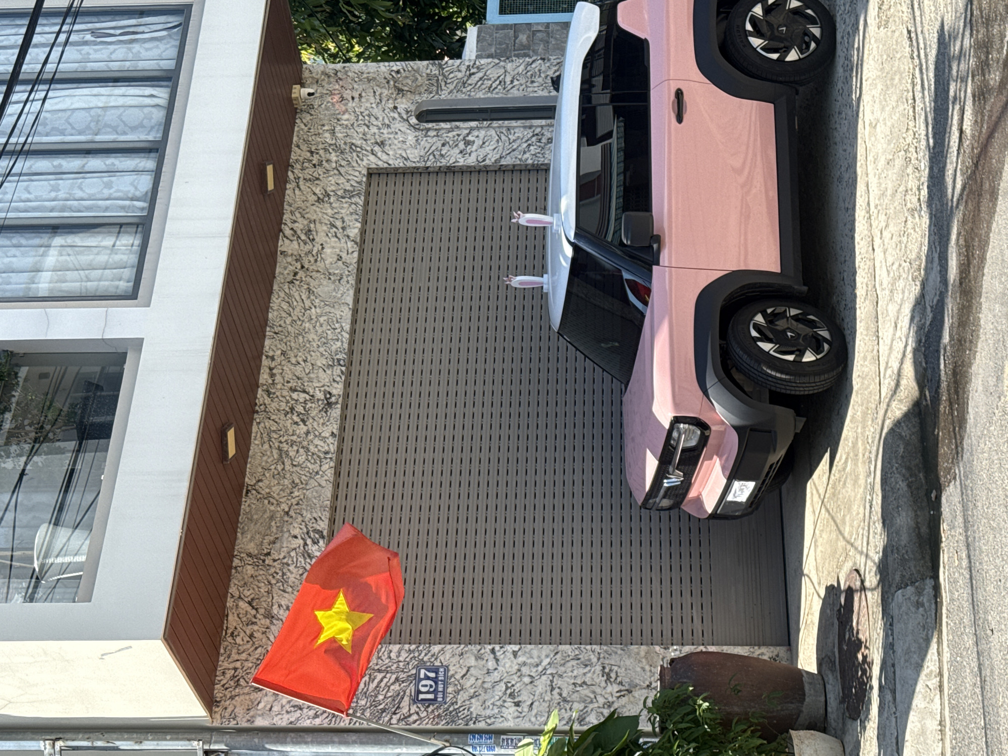 In Bac Nha Trang Ward, Vietnam, a pink car with white accents and bunny ears on its roof is parked in front of a house. The house has a gray, textured facade and a large, gray roller garage door. A Vietnamese flag flies to the left of the garage door. To the left of the flag, a house number "197" is visible on a blue sign, with the street name "BUI HUY DICH" below it. A plant in a large pot sits on the left side of the image. The scene is illuminated by sunlight, casting shadows on the ground.