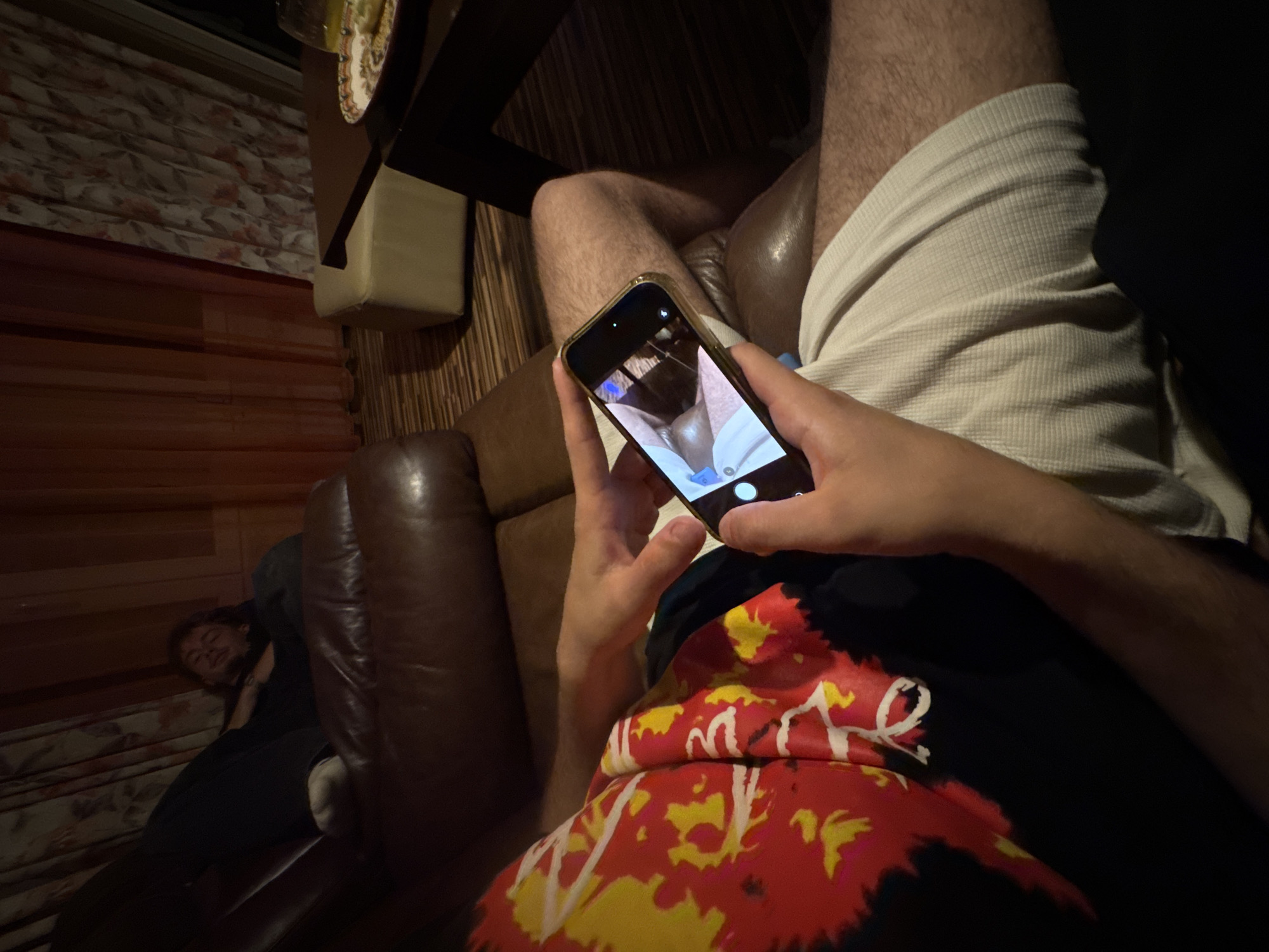 The scene depicts an indoor setting, identified as being in the city of Sofia, Bulgaria. Two individuals are present amidst residential-style furniture.
In the foreground, a person is partially visible from the chest down, seated on a brown leather sofa. This individual wears a black top with prominent red, yellow, and white abstract patterns, and light-colored, possibly off-white, textured shorts. Their bare, hairy legs are visible. The person holds a gold-colored smartphone horizontally with both hands, its screen displaying a camera application interface with a prominent white circular shutter button. The phone's camera view appears to be directed towards the background.
In the mid-ground, a second individual is fully reclined on a separate brown leather sofa. This person wears dark clothing and has dark hair, presenting a relaxed posture with a faint smile or smirk on their face. One foot, encased in a white sock, is propped on the sofa armrest.
The environment includes multiple pieces of furniture: two brown leather sofas, a light-colored upholstered ottoman, and dark wooden tables. On one table, a colorful, patterned plate holds food items (resembling pastries or snacks) and a glass contains a yellowish beverage. The room features patterned curtains, one with a floral design and another in a solid peach or orange hue, along with a dark, wood-patterned floor. The overall lighting is dim.