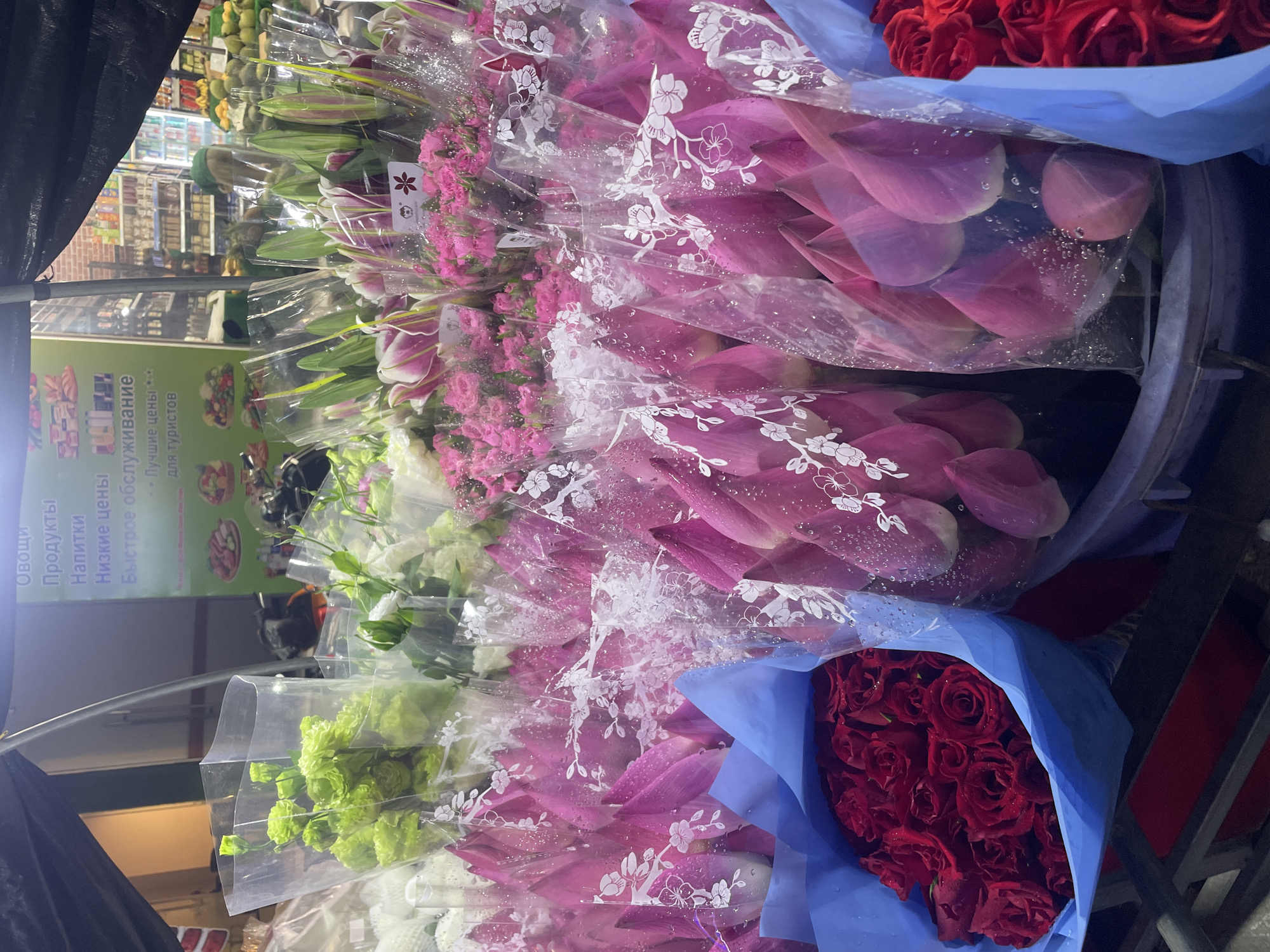 The image captures a vibrant display of flowers at what appears to be a market stall or storefront in Bắc Nha Trang, Vietnam, likely catering to tourists. The view is predominantly filled with numerous bouquets arranged vertically, creating a lush, colorful wall.
The main subjects are various types of flowers, individually wrapped and bundled. Prominently featured are large bouquets of red roses, wrapped in bright blue paper, positioned towards the right side of the frame. Adjacent to them, and spanning much of the central and upper-left area, are numerous bundles of what appear to be pink lilies or similar large-petaled flowers, encased in clear plastic wrap adorned with white floral patterns. Many of these plastic wraps show water droplets, suggesting freshness or recent misting. Towards the lower and upper-left, there are also bundles of green and white flowers, possibly eustoma or carnations, also wrapped in clear plastic.
In the background, visible through gaps in the flower display, are shelves stocked with various grocery items, including fruits and packaged goods. The setting suggests an indoor or sheltered outdoor market environment, illuminated by artificial lighting, indicating it could be evening or night.
A notable detail is a green sign with white text in Russian, which reads: "Овощи" (Vegetables), "Продукты" (Groceries), "Напитки" (Drinks), "Низкие цены" (Low prices), "Быстрое обслуживание" (Fast service), and "- Лучшие цены для туристов" (Best prices for tourists). This sign strongly indicates that the establishment targets a Russian-speaking tourist demographic. The overall scene conveys a bustling commercial atmosphere centered around the sale of fresh produce and flowers.