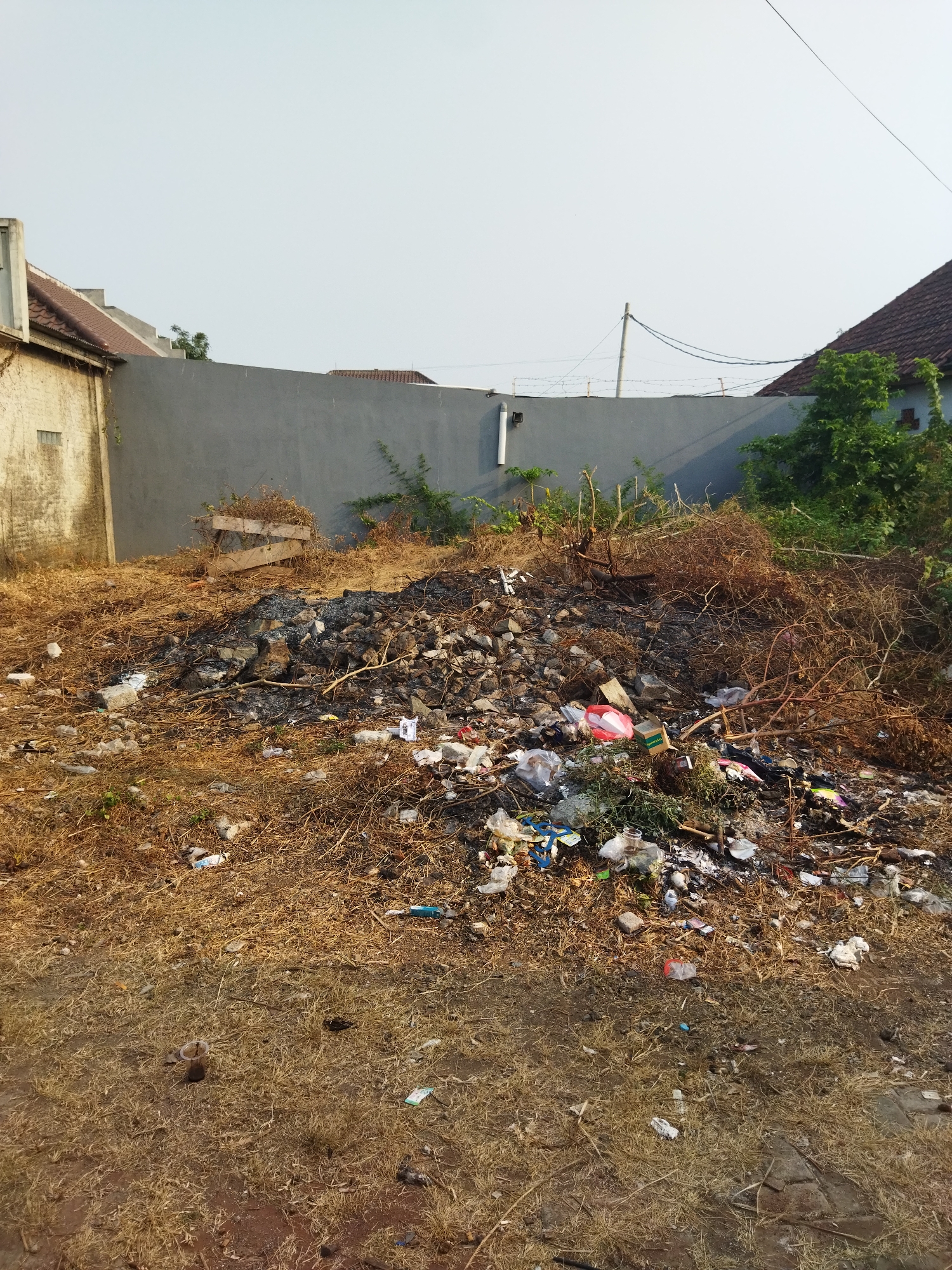 The image depicts an outdoor scene of accumulated debris and vegetation. In the foreground, dry, brown grass covers the ground, interspersed with scattered pieces of litter. A pile of dark, burnt material, likely the remnants of a fire, sits centrally. This pile is surrounded by a mix of dry branches, leaves, and more scattered trash, including plastic bags, wrappers, and what appears to be a red plastic container.
Behind this debris, a grey, concrete wall forms a backdrop. To the left of the wall, a section of a building with a light-colored textured wall and a tiled roof is visible. To the right of the wall, dense green foliage and trees obscure parts of the residential structures, which have dark tiled roofs. Overhead, a clear sky is visible, with power lines and a utility pole present. The overall impression is one of neglect and waste accumulation in an urban or semi-urban setting. The location is identified as Suka Sari, Indonesia.