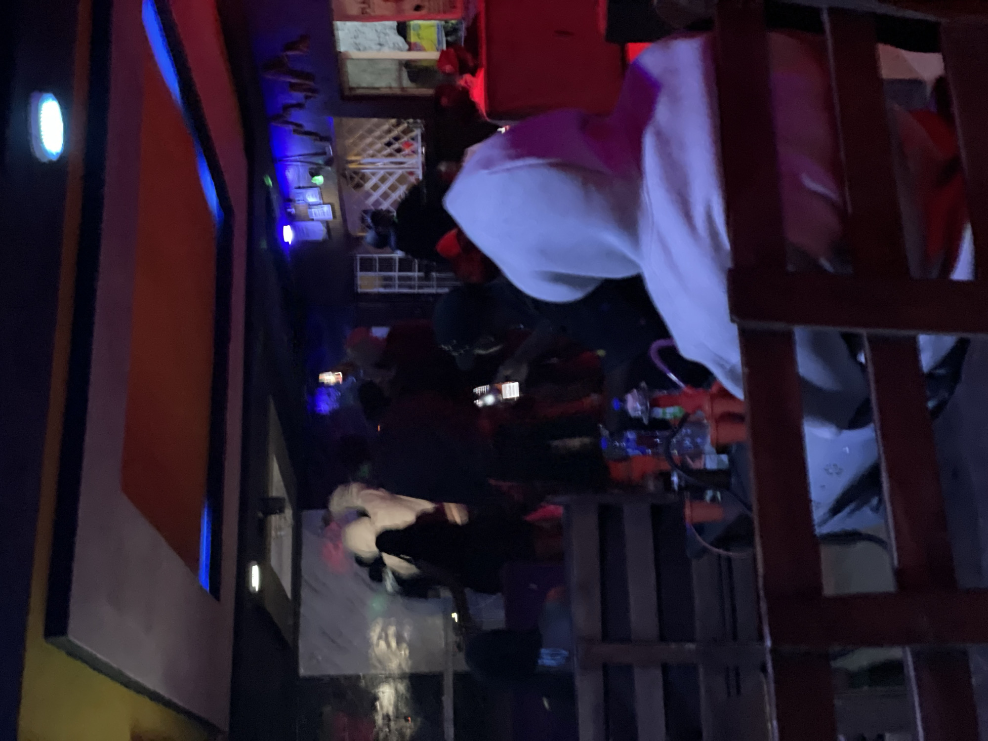 The image shows the interior of a dimly lit establishment, possibly a bar or lounge, located in Durumi, Nigeria. The space is characterized by a modern ceiling design with recessed lighting, featuring blue and orange elements. Several individuals are present, mostly silhouettes due to the low lighting and the angle of the photograph. In the foreground, a person wearing a white hooded sweatshirt is seated facing away from the camera, their back and head visible. Behind them, other patrons are gathered, some standing and some seated at tables or along a wooden bar. The atmosphere appears social, with groups of people interacting. Drinks and possibly shisha pipes are visible on surfaces in the midground. The overall impression is one of a relaxed, evening gathering in an urban setting.
