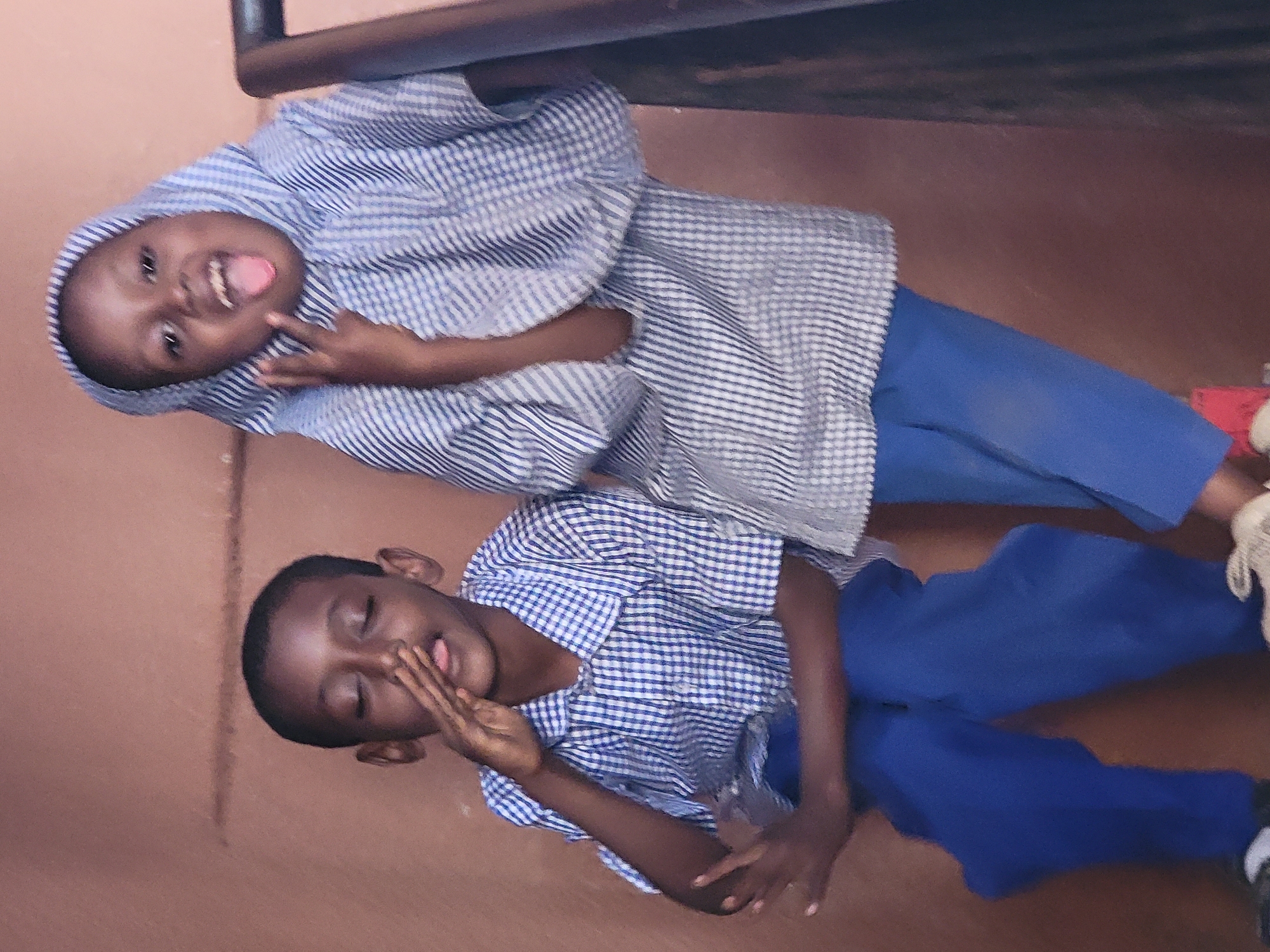 This image, taken in Maiburiji, Nigeria, features two children against a plain reddish-brown background. The photograph is oriented vertically (rotated 90 degrees clockwise), depicting the children in what appears to be playful standing poses. Both children have dark skin tones and are dressed in similar blue trousers and blue and white patterned tops, suggestive of school uniforms.
The child positioned higher in the frame wears a light blue and white striped long-sleeved top, which includes a hood or head covering. This child smiles broadly at the camera, sticking out their tongue, and holding up two fingers in a gesture often associated with a peace sign or "V" sign.
The child below them wears a short-sleeved blue and white checkered shirt, paired with blue trousers. This child has their eyes closed and is also sticking out their tongue, while their hands are pressed together covering part of their mouth in a gesture that seems playful or slightly shy.
The scene conveys a cheerful and spontaneous moment. The children's expressions indicate happiness and a lighthearted interaction with the camera. The background is minimalistic, suggesting an indoor setting or a plain wall. A dark, horizontal bar or ledge runs across the top of the frame. No text is visible in the image. The lighting is consistent and diffuse, without harsh shadows.