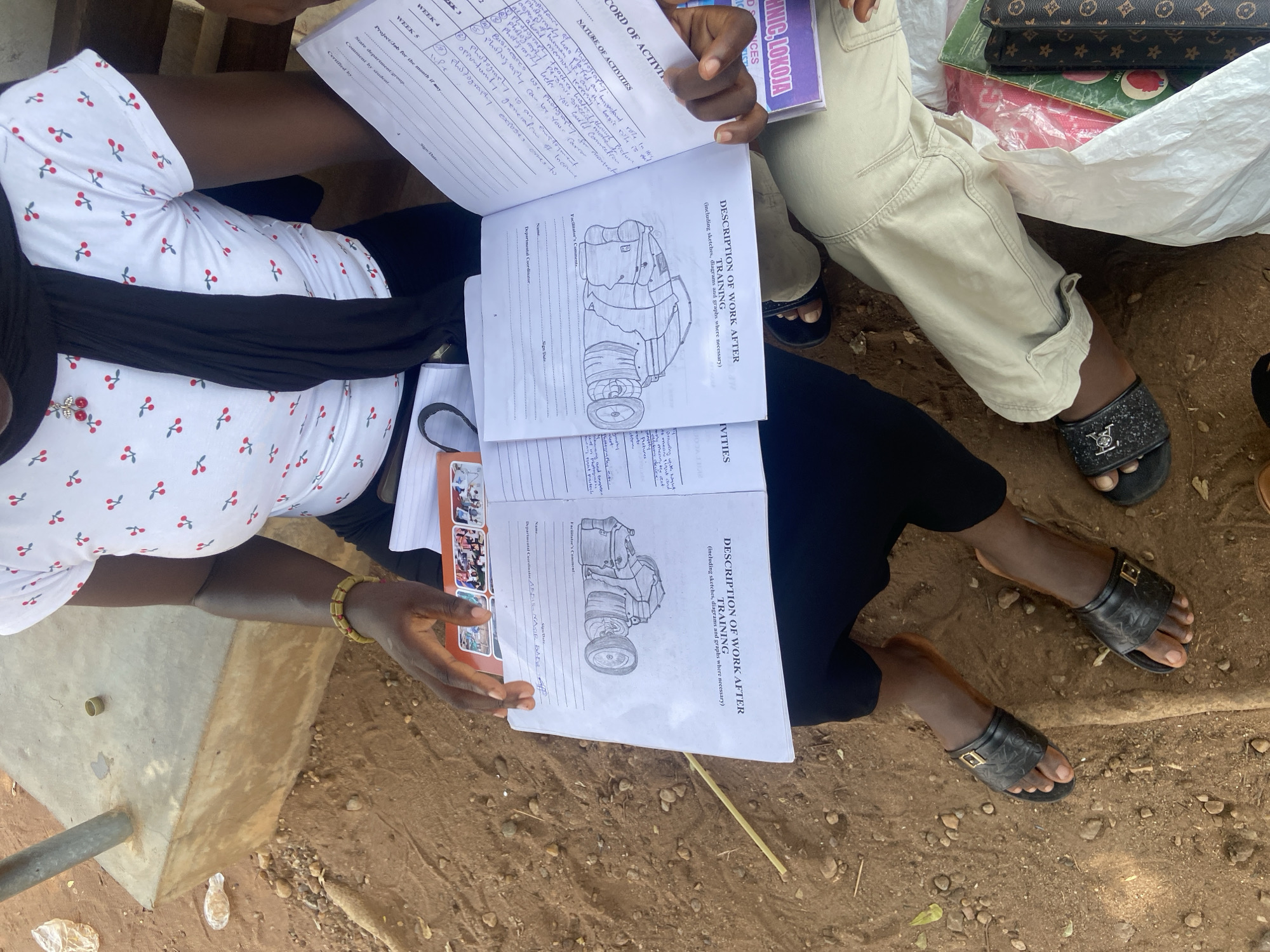 Here is a detailed description of the image:
**Overview:**
The image presents an overhead view of two individuals engrossed in examining documents, presumably related to a training program.
**People:**
* Two individuals are visible. One is wearing a white top with a cherry pattern and a black head covering. The second person is wearing beige pants. Both are sitting.
**Subjects:**
* The main focus is on the open books or reports being held and read by the individuals. They have titles and diagrams on them. The documents pertain to "Record of Activities" and "Description of Work After Training."
**Objects:**
* Books/Documents: The primary objects are the open documents with text, diagrams, and charts.
* Clothing: Details include the cherry-patterned white top, black head covering, beige pants, and sandals worn by both individuals.
* Ground: The background appears to be a patch of dry, reddish-brown soil, possibly a dirt yard.
**Actions/Interactions:**
* The individuals are engaged in reading and reviewing the documents.
**Environment:**
* The scene appears to be outdoors.
**Location:**
Based on the information in the image the location is Lokoja, Nigeria.