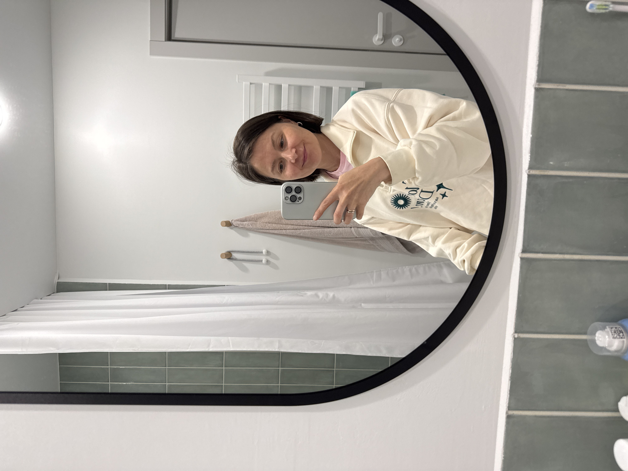 A woman is taking a selfie in a bathroom mirror. She is holding a smartphone in her right hand, which is partially obscuring her face. She is wearing a light-colored zippered sweatshirt over a pink shirt. Behind her in the mirror's reflection is a white shower curtain, green tiles, a towel rack with a towel hanging on it, and a white towel warmer. A toothbrush is also partially visible in the lower right corner of the image, placed on the tiled surface. The mirror has a black round frame. The specific location in Izhevsk, Russia, cannot be determined from the visual information provided.