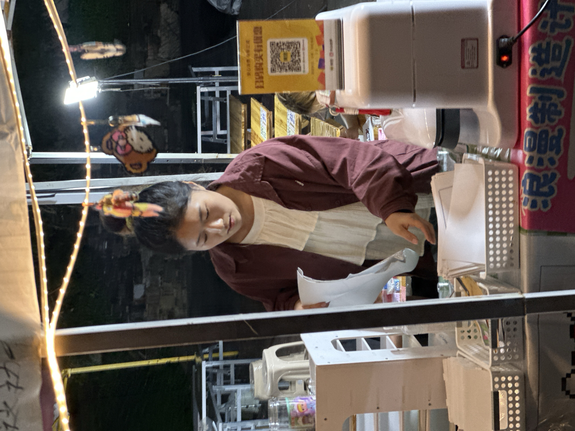 A young Asian woman with her hair pulled back in a bun, wearing a maroon jacket over a white ruffled blouse, is working at a market stall at night. The stall is set up under an awning strung with decorative lights. She is holding a sheet of paper and appears to be attending to a customer or preparing goods. To her right, a yellow sign with a QR code and Chinese text is prominently displayed on top of a white appliance, likely a payment terminal or a device for a service. The stall is filled with various items, including white baskets containing what appear to be supplies or merchandise. In the background, there are wooden tables and other market stalls, suggesting a bustling night market environment. The location is identified as the Chongqing High-tech Zone, China.