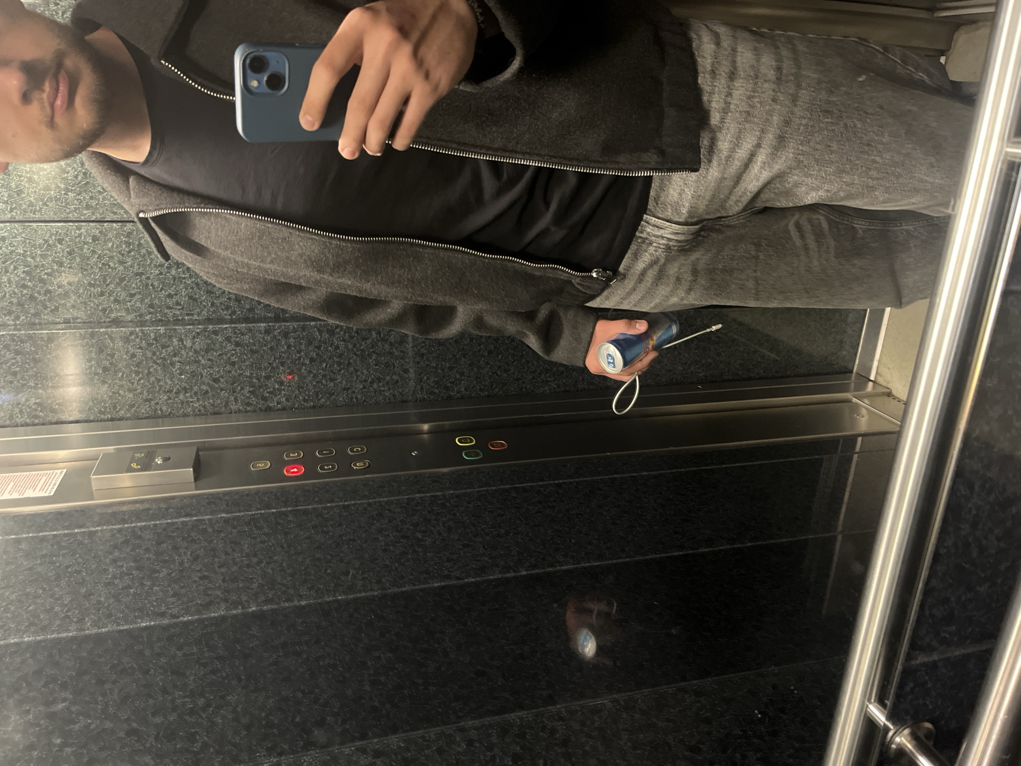 The photo shows a man in an elevator. He is holding a blue smartphone in his left hand, likely taking a selfie. He is wearing a black t-shirt under a gray jacket and gray jeans. In his right hand, he holds a can of Red Bull with attached earphones. The elevator has buttons for floors 1-5, as well as ground level and basement levels. The interior of the elevator is finished in dark, textured panels, possibly with stainless steel accents and handrails. The location of the photo is not possible to determine based on the image, and as such, the city of Blagoevgrad, Bulgaria, cannot be inferred.
