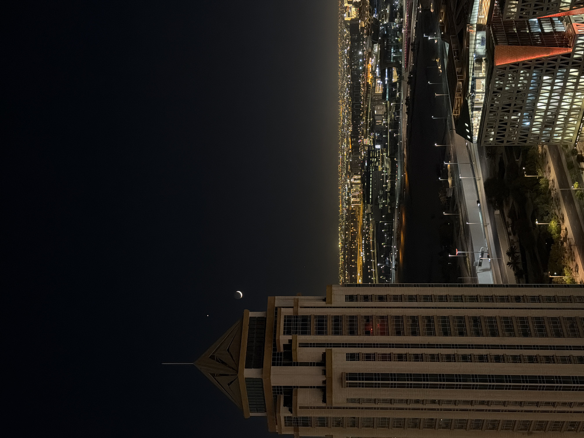 An aerial nighttime, vertical, medium shot captures a crescent moon and a bright star in the dark sky above a sprawling cityscape in Unknown, Unknown. In the foreground, a tall, beige, multi-story building with numerous windows dominates the left side of the frame. Its sharp, angular roof is capped with a thin, white antenna. To the right, a lower, modern building with a distinctive angular facade, illuminated with warm orange and white lights, is visible. In the background, the city extends to the horizon, a dense tapestry of lights from buildings and streetlamps. A highway with passing cars runs through the middle ground, bordered by streetlights and scattered trees.