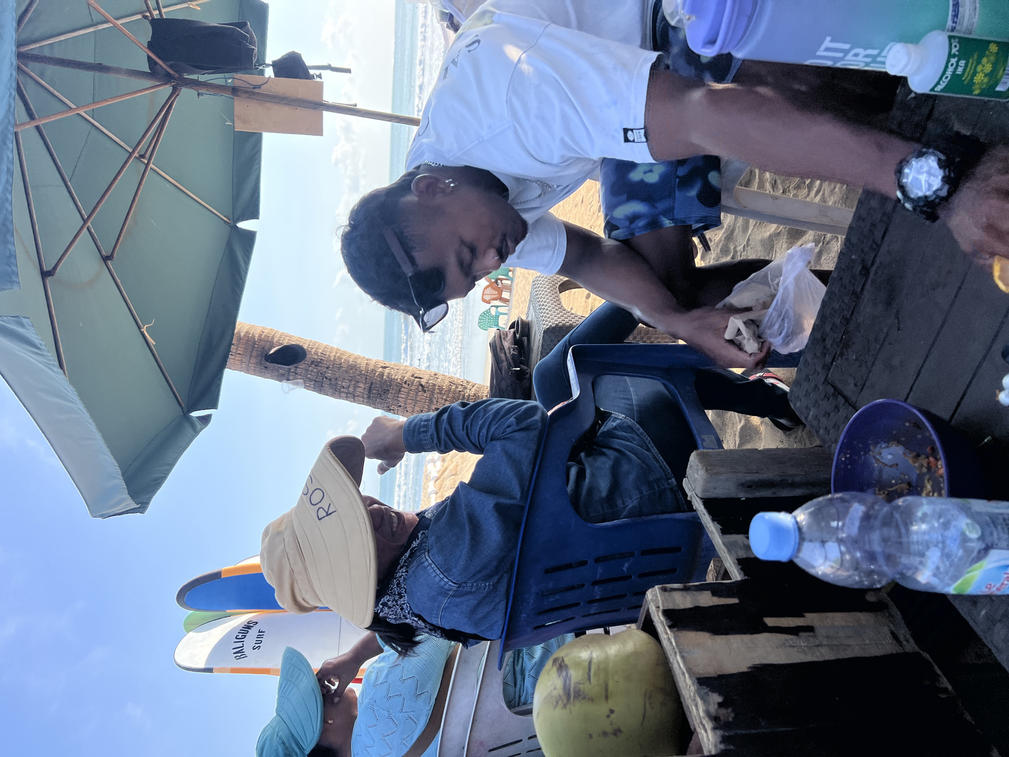 This image captures a scene on a beach in Kuta, Indonesia, likely near a surf spot given the surfboards in the background. Two people are seated at a table, with a third person partially visible in the background. In the foreground, a man wearing a white t-shirt and patterned shorts is looking down at something in a plastic bag he is holding. He has sunglasses pushed up on his head and is wearing a watch. To his left, a woman in a denim jacket and a wide-brimmed hat that reads "ROSE" is seated in a blue plastic chair, smiling and looking towards the left. To her left, another person wearing a blue top and a light blue hat is visible. Behind them, several surfboards are propped up, two of which have "BALIGUNS SURF" written on them. A large green umbrella provides shade over the seating area. On the table, there is a clear plastic bottle of water, a purple bowl with some food remnants, and a green coconut. The background shows the ocean with gentle waves and a sandy beach with a few colorful chairs. The overall atmosphere suggests a relaxed, casual setting on a sunny day.