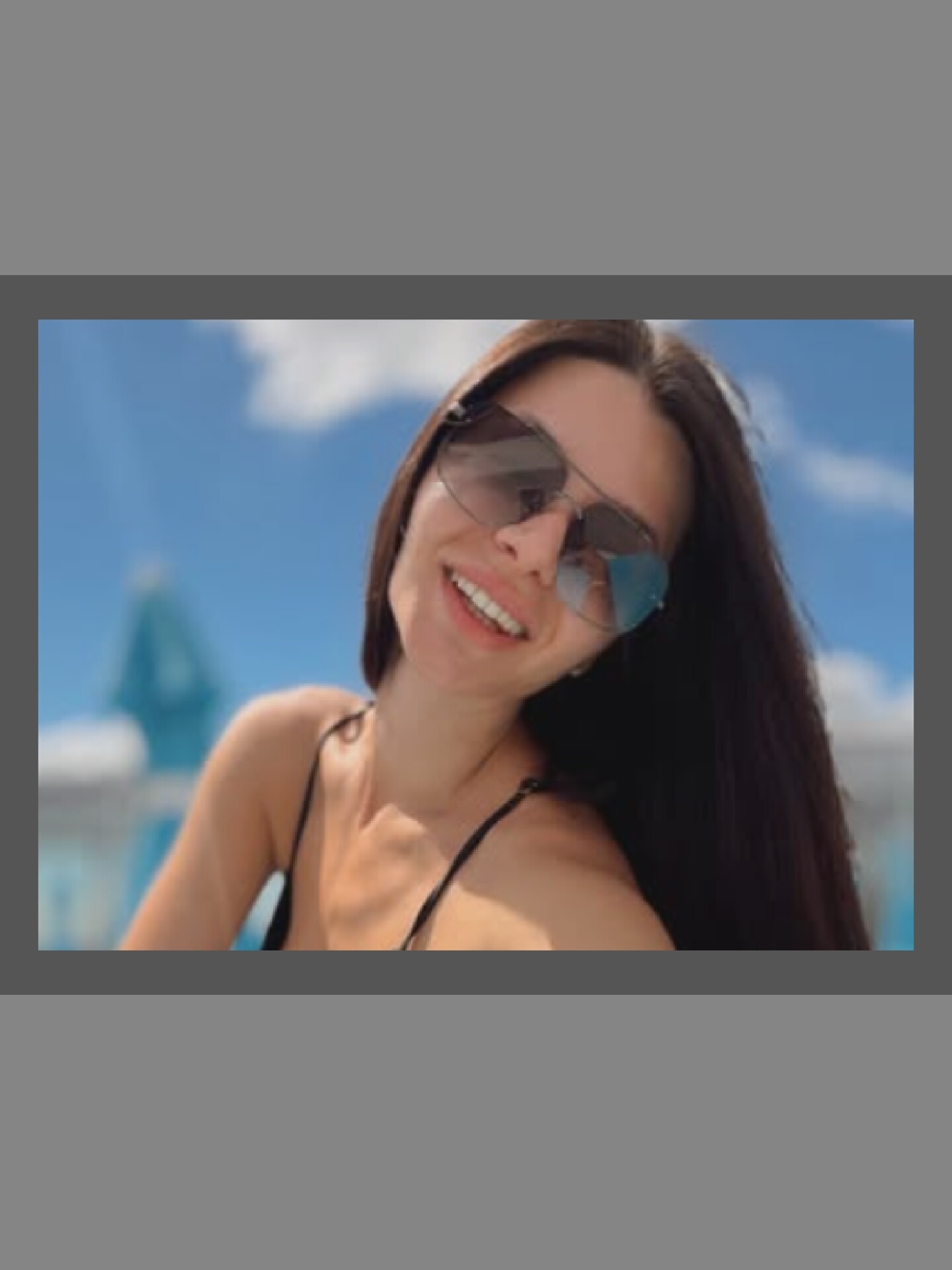 A young woman with dark hair and sunglasses smiles at the camera in a close-up shot. She is wearing a black bikini top. The background is blurred and shows a bright blue sky with some clouds, and what appears to be a swimming pool area with a blue structure. The lighting suggests it is daytime and sunny.