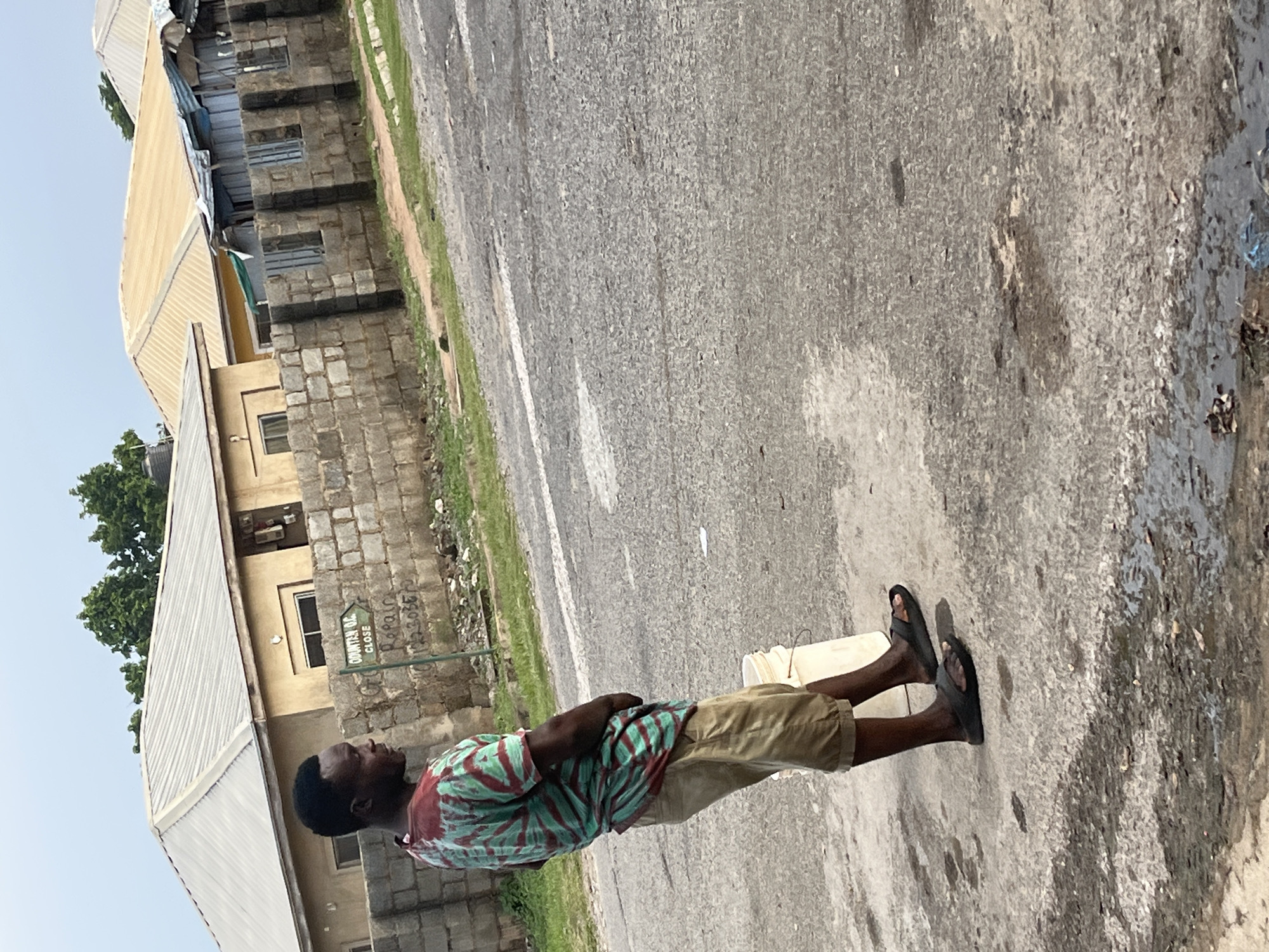 A dark-skinned man stands on a paved road, facing to the right and looking away from the camera. He is wearing a green and red tie-dye t-shirt and khaki shorts, with black flip-flops on his feet. A white bucket is positioned to his right. In the background, a low wall made of concrete blocks separates the road from a residential area with buildings featuring corrugated metal roofs. A green sign with white text indicating directions is mounted on a post to the left of the man, reading "COUNTER CE CLOSE" followed by a series of numbers. The environment appears to be an outdoor urban setting.