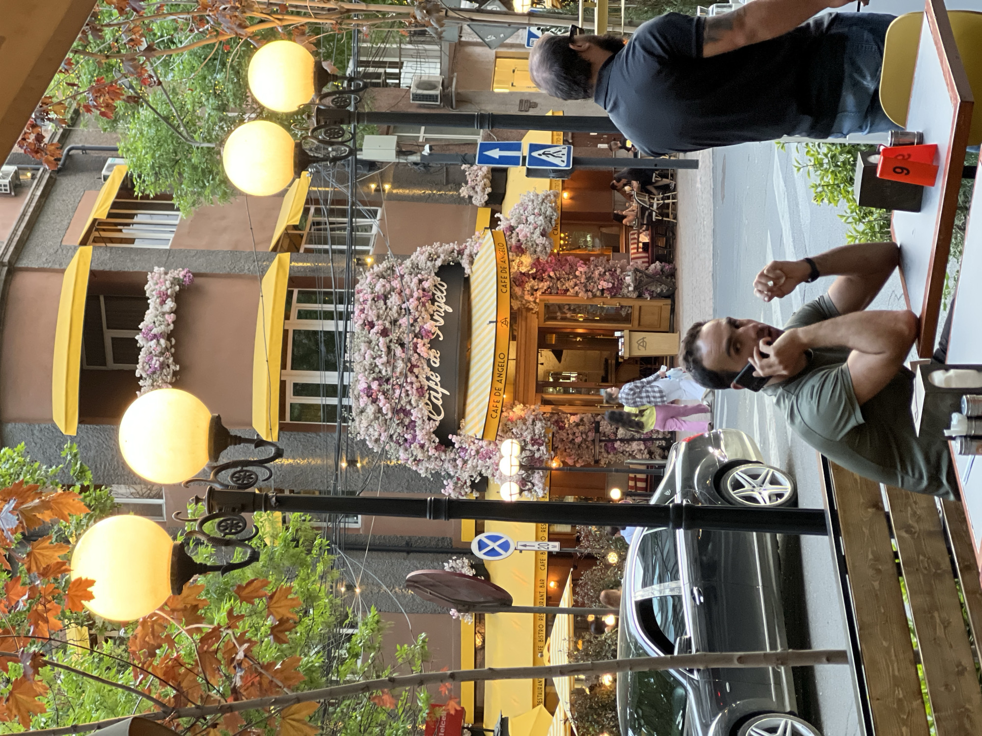 Here is a forensic description of the image:
**Setting:** An outdoor scene at Cafe de Angelo, a cafe in Yerevan, Armenia. The cafe's exterior is decorated with flowers. Street signs are present.
**Objects:** The scene includes street lights, a wooden bench, a table with a red number "9" stand, a car, a restaurant, and street signs.
**People:** Two men are at a table, one is using a mobile phone. Another individual is further down the sidewalk.
**Environment:** The weather appears to be daytime. Autumn leaves are on the tree on the left side of the image.