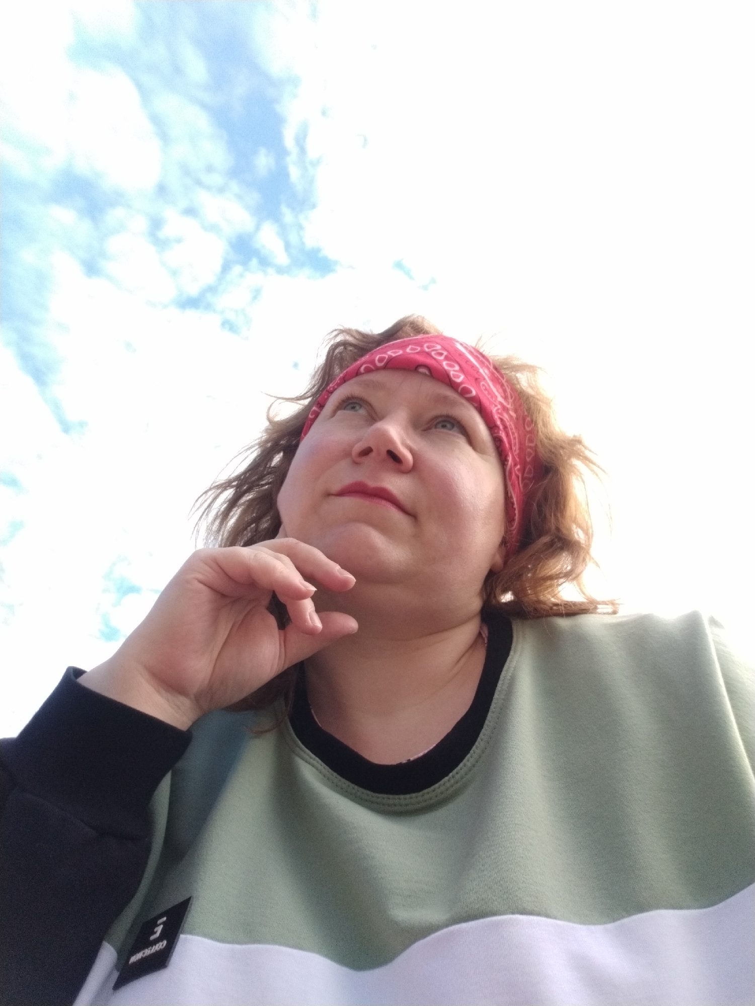 A woman wearing a red bandana and a sweatshirt with white and green panels is looking upwards with her chin resting on her hand. The sky, visible in the background, is blue with scattered clouds. The location is unknown.