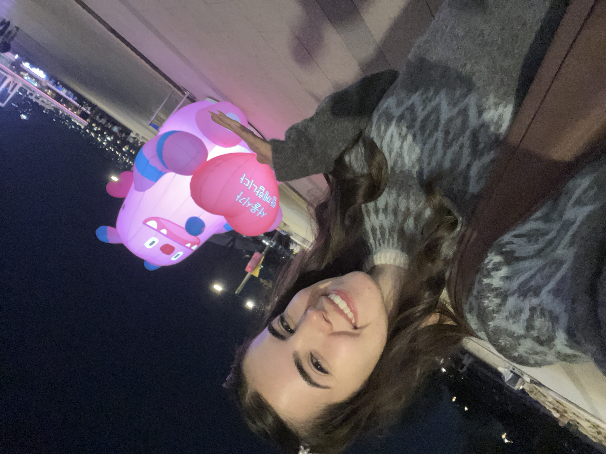 A woman is taking a selfie at night. In the background is a large, inflated pink cartoon character. The cartoon character is a hippo with big eyes, open mouth and arms out. It has pink and blue spots, and a pink heart balloon with Korean script. The background appears to be a city skyline at night, likely in Seoul, South Korea due to the script on the balloon. The woman has long brown hair and is wearing a gray and white patterned sweater.