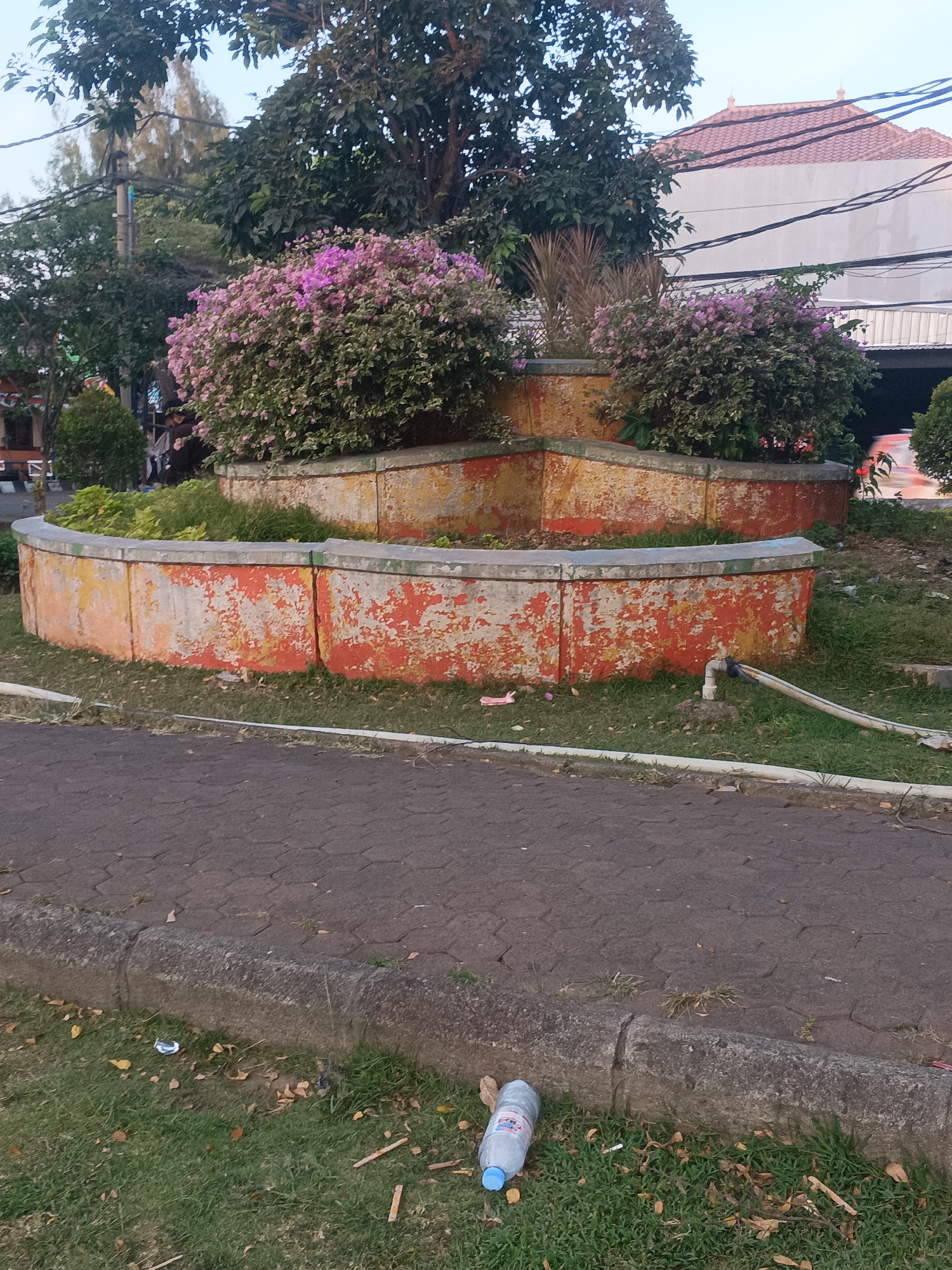 The image depicts an outdoor scene, likely a public park or garden area in Surabaya, Indonesia. In the foreground, a paved walkway made of interlocking bricks runs parallel to a concrete curb bordering a grassy area. Scattered on the grass and along the curb are fallen leaves and a plastic water bottle with a blue cap lying on its side.
Dominating the midground is a tiered, circular planter box constructed of weathered concrete. The exterior of the planter is painted with peeling layers of orange and yellow. Within the planter, lush green foliage and clusters of vibrant pink bougainvillea flowers are visible. Behind the planter, trees with dense green leaves form a backdrop, and a portion of a building with a tiled roof can be seen in the upper right of the frame, along with overhead power lines. The overall impression is a somewhat neglected urban green space.