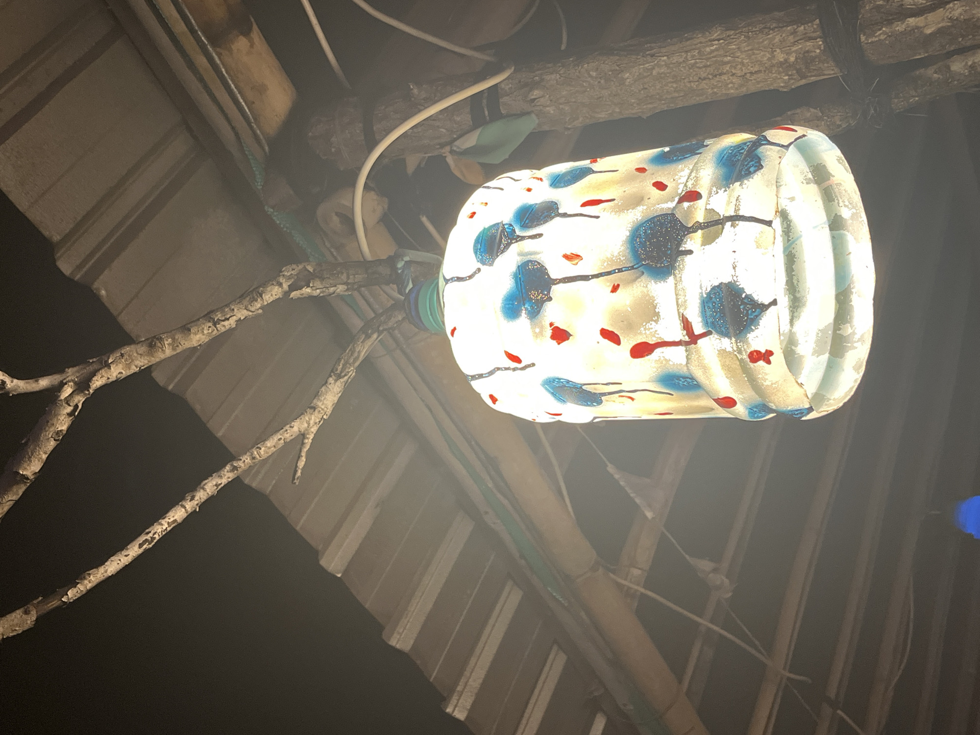 This image captures a close-up view of a homemade, illuminated lantern at night, set against a rustic background. The central subject is a horizontally oriented, cylindrical lamp, appearing to be crafted from a plastic bottle. It is decorated with an abstract pattern of blue and red paint splatters or dots on a translucent, light-colored surface. The lamp emits a bright, warm white light, suggesting an internal bulb.
The lamp is integrated into an improvised structure, supported by and surrounded by natural, textured tree branches or twigs. These branches crisscross the frame, some appearing to be part of the supporting framework for the lamp or overhead structure. Various electrical wires and cables, some white and some darker, are visible intertwined with the branches, hinting at an outdoor or semi-outdoor electrical setup.
In the background, dark, corrugated metal sheets are discernible, likely forming part of a roof or wall. Some bamboo-like poles are also visible. The overall setting is dark, indicating nighttime, with the lamp serving as the primary light source, creating a warm ambiance. A faint blue light source, out of focus, is barely visible in the lower right corner, suggesting additional decorative lighting. There are no people visible, and no specific activity is taking place other than the lamp providing illumination. The scene conveys a simple, possibly improvised, and functional lighting arrangement in an unspecified location, consistent with the provided context of "Unknown, Indonesia." No visible text is present in the image.