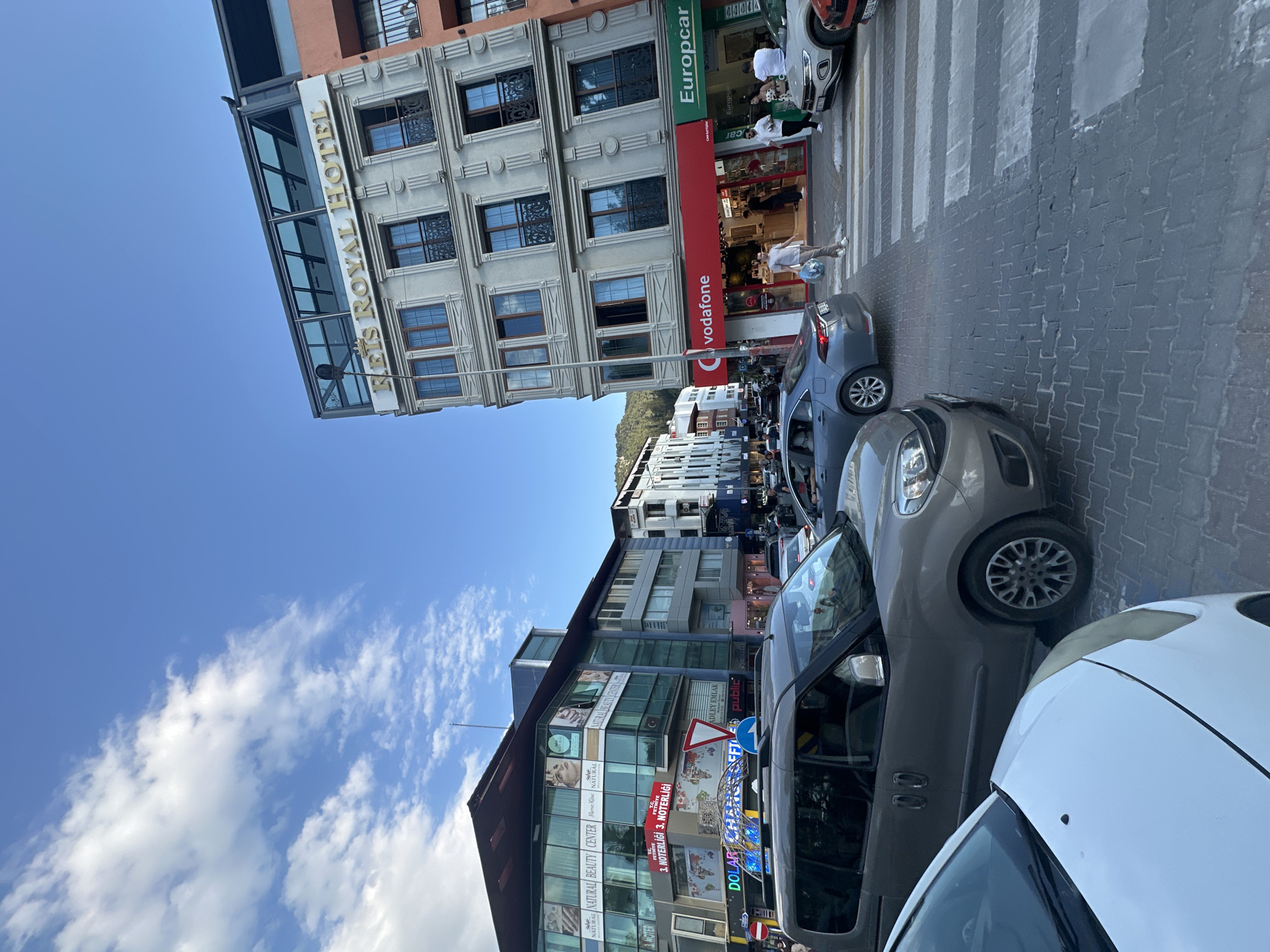 The image depicts a street scene in Fethiye, Turkey. The "REIS ROYAL HOTEL" is visible with a glass extension on the side. A pedestrian crossing is present with a person walking across it. Various shops are visible, including Vodafone and Europcar. Several cars are parked along the street. The upper left portion of the image shows a blue sky with clouds. Other buildings, including one housing a "NATURAL BEAUTY CENTER" and "3. NOTERLIGI" (3rd Notary) are also observable. Street signs and advertising are prominent.