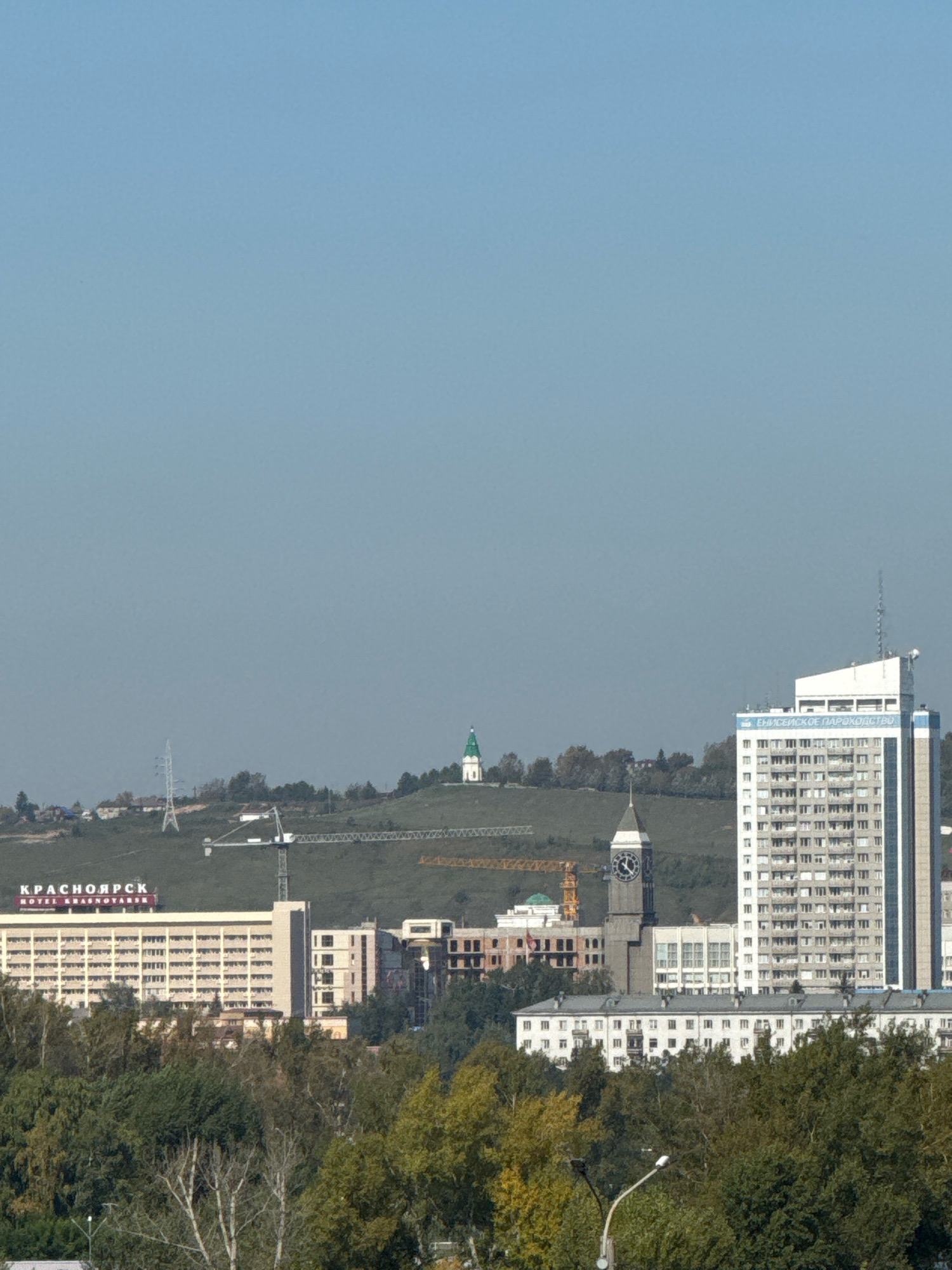 This is a daytime, eye-level, outdoor, medium shot of a cityscape in Krasnoyarsk, Russia. In the foreground, a dense canopy of green and yellow foliage dominates the bottom of the frame, with a street light pole visible on the left. Beyond the trees, a long, low-rise building with a white facade and multiple windows stretches horizontally. To the left of this building, a larger, multi-story hotel is visible, displaying the Cyrillic text "КРАСНОЯРСК" and "HOTEL KRASNOYARSK" in red lettering.
Further in the background, a prominent clock tower with a dark roof and a large clock face stands tall. To its right, a tall, white and blue high-rise building features numerous windows and a satellite dish on its roof. The text "ЕНИСЕЙСКОЕ ПАРОХОДСТВО" is visible in blue on the side of this building.
On a distant, verdant hill, a white building with a green dome, likely a church, is visible. A large construction crane with a yellow boom is positioned on the hillside, suggesting ongoing development. A tall power line pylon is also present on the hill. The sky above is clear and pale blue. The overall scene depicts a mix of older and modern architecture, green spaces, and signs of urban development within the city.