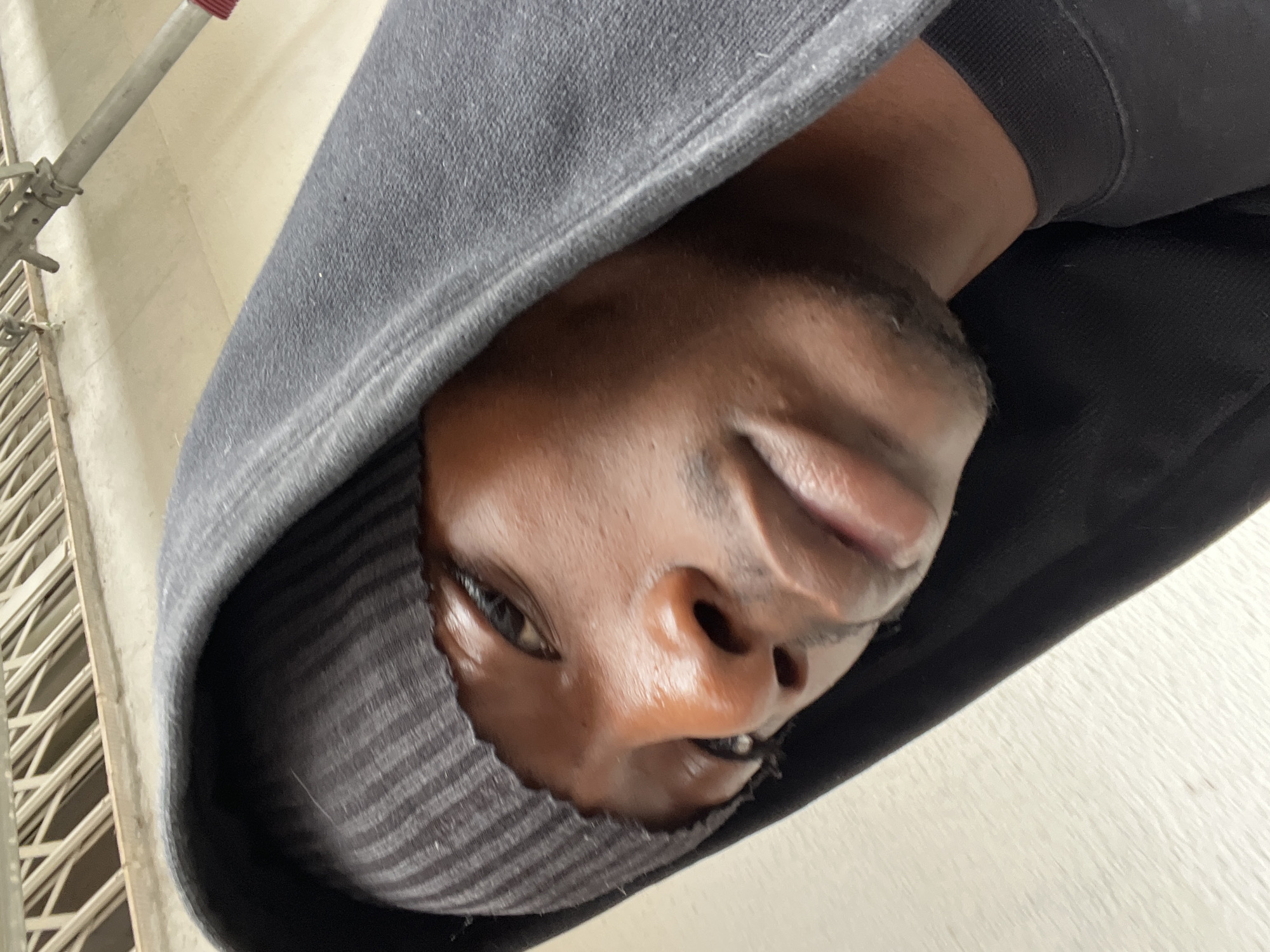 An upward-angled close-up of a dark-skinned person wearing a dark-colored hooded sweatshirt. The person's face is the focal point, with the hood partially obscuring the forehead and neck. The individual has dark eyes, a prominent nose, and full lips. The person is wearing what appears to be a black metal piercing in the bottom lip. The hood appears to be two layers or is turned over. Behind the person, on the left side of the frame, is a blurred vertical structure that resembles scaffolding or a metal grate. The background appears to be a light-colored wall or surface. There is no information to conclude location of Barcelona, Spain.