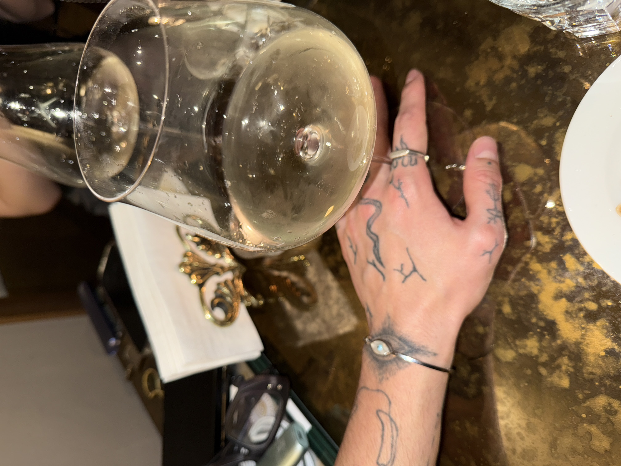 A table setting in what appears to be a restaurant in Yerevan, Armenia. The table is a dark golden color with a slightly reflective surface. A hand with multiple tattoos and rings is placed on the table. There is also a silver bracelet with an oval stone on the wrist. Two wine glasses, filled with a light-colored liquid, are partially visible. A white plate is in the upper right corner, and a pair of eyeglasses and a white napkin with gold embellishments are also visible. The lighting is generally bright, suggesting daylight or well-lit interior conditions.