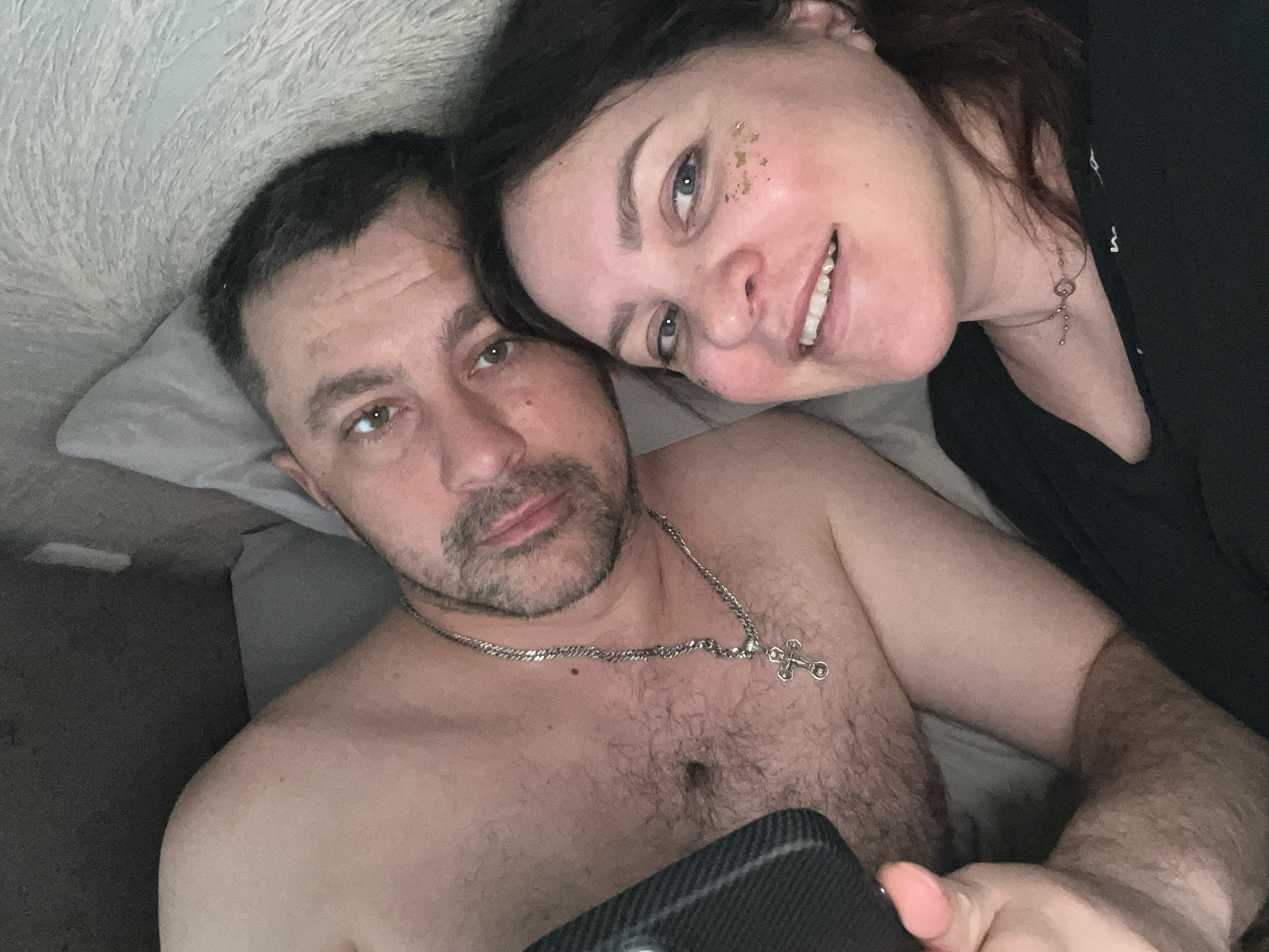 A close-up selfie depicts a man and a woman in what appears to be a bed. The man, shirtless and with a silver chain and cross pendant, lies with his head on a pillow. He has short, dark hair, stubble, and a serious expression. The woman leans her head toward his, smiling and looking into the camera. She has dark hair, fair skin, and what seems to be decorative gold glitter on her cheek. She wears a black garment, partially visible. The background is indistinct, suggesting an indoor setting. There's no indication this image was taken in Noviy, Russia. The location cannot be verified. The man holds a smartphone.