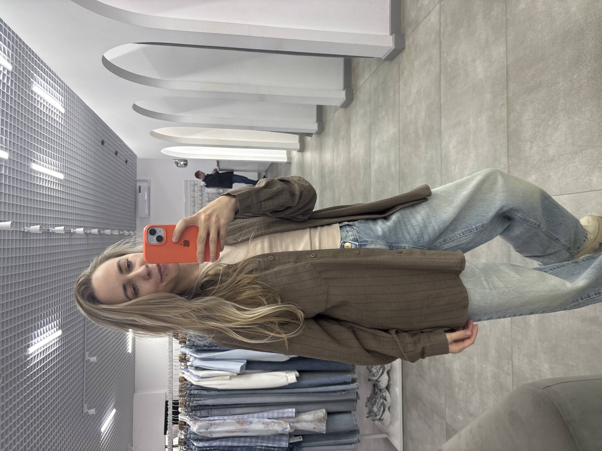 A blonde woman is taking a selfie in a clothing store. She is wearing light blue jeans, a light-colored t-shirt, and a brown, button-down shirt. She is holding an orange smartphone in her left hand. Behind her, there is a rack of jeans. In the background, there are dressing rooms with arched entrances. The floor is tiled in gray. The ceiling has a grid pattern with rows of lights. In the far background, a person is standing in one of the dressing room entrances. The store is located in Chaikovsky, Russia.