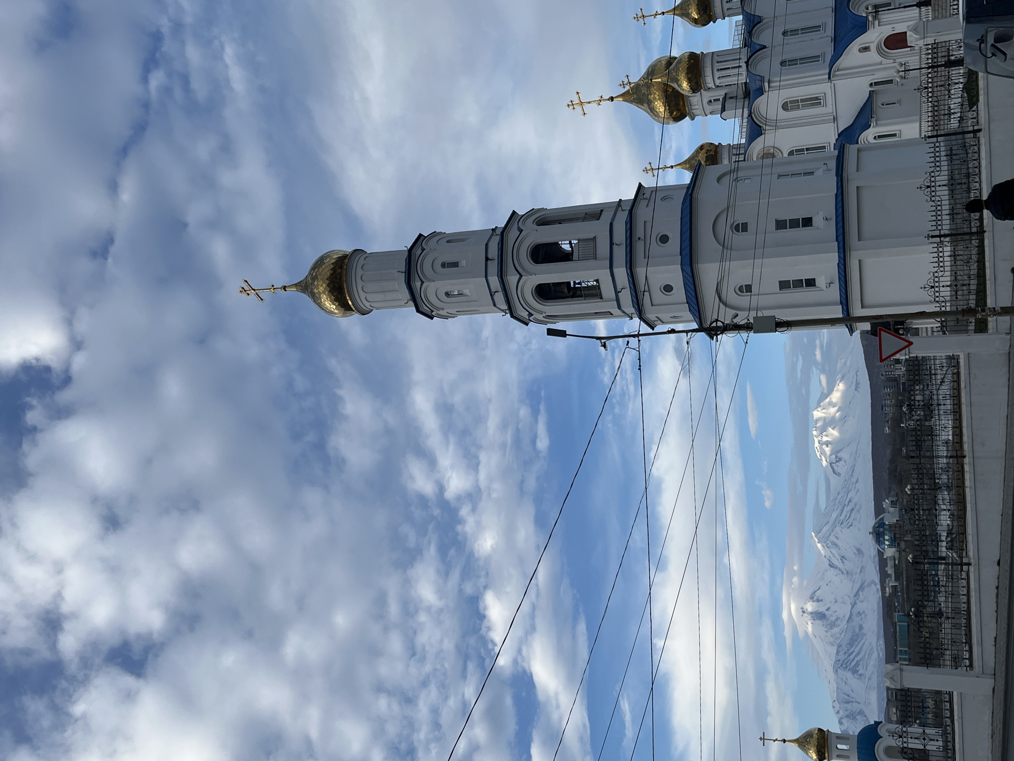 The image depicts an upward-facing view of a Russian Orthodox church with a tall, white bell tower and golden domes against a cloudy blue sky. The church features white walls with blue accents on the roof edges. A series of golden domes with crosses top the structures. In the background, snow-capped mountains are visible, suggesting a landscape with mountainous terrain. The scene is an urban environment, with buildings and a metal fence visible. Electrical wires are strung across the foreground of the sky. A triangular traffic sign can be seen on the right side of the image. A person is faintly visible in the right side. The location is Petropavlovsk-Kamchatsky, Russia.