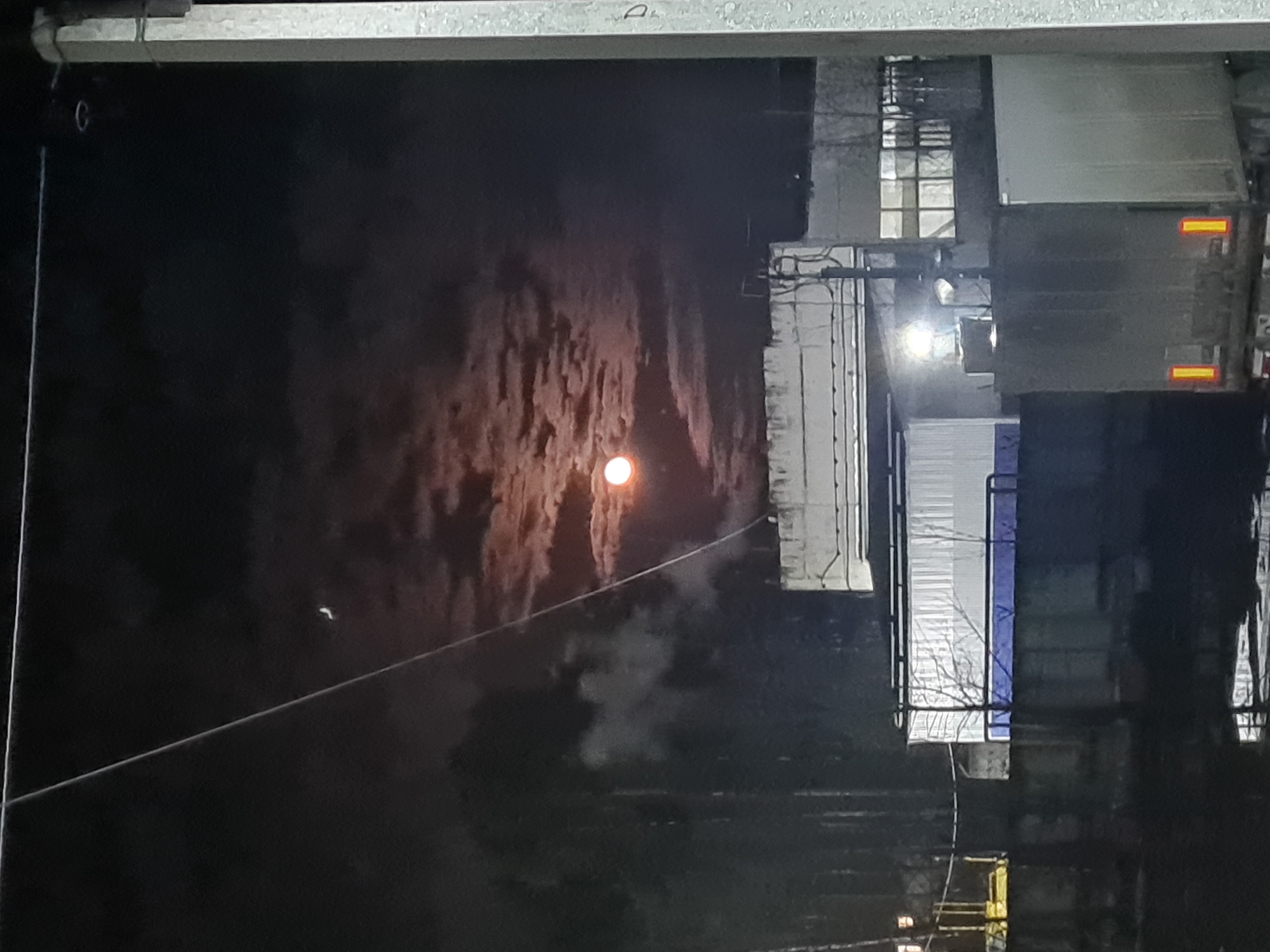 The image depicts an outdoor scene at night. A bright, circular light source, possibly the moon, is visible in the left side of the frame, surrounded by dark, textured clouds. A thin wire or cable stretches diagonally across the lower left quadrant of the frame. On the right side, there's an array of structures, seemingly buildings or industrial constructions, including what appears to be a truck or large vehicle with visible lights and reflective markers. Some of the buildings have lit windows. The image is taken from an elevated position, possibly from a window or balcony, and the scene suggests an urban or industrial environment. The location is likely Noviy, Russia. There are no visible people or specific actions taking place. The overall environment is dark and overcast.