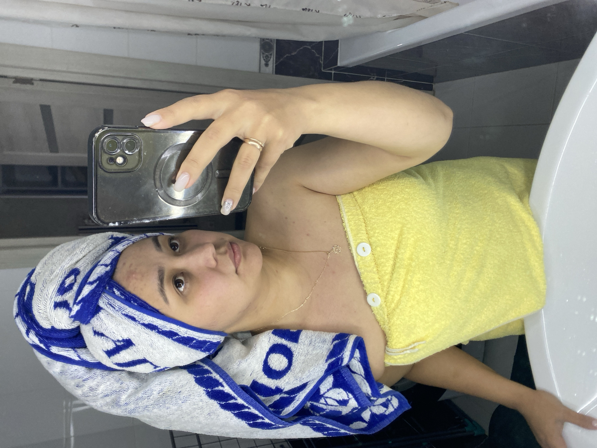 A woman in a bathroom takes a selfie in a mirror. She is wearing a blue and white towel turban on her head and a yellow towel wrap around her body. She is also wearing a delicate gold necklace with a flower pendant. In her right hand, she holds a smartphone, displaying a silver phone case. A gold ring adorns her ring finger. The background reveals a tiled wall, a bathtub, and a door. The scene suggests a domestic setting, possibly her home bathroom in Ufa, Russia.