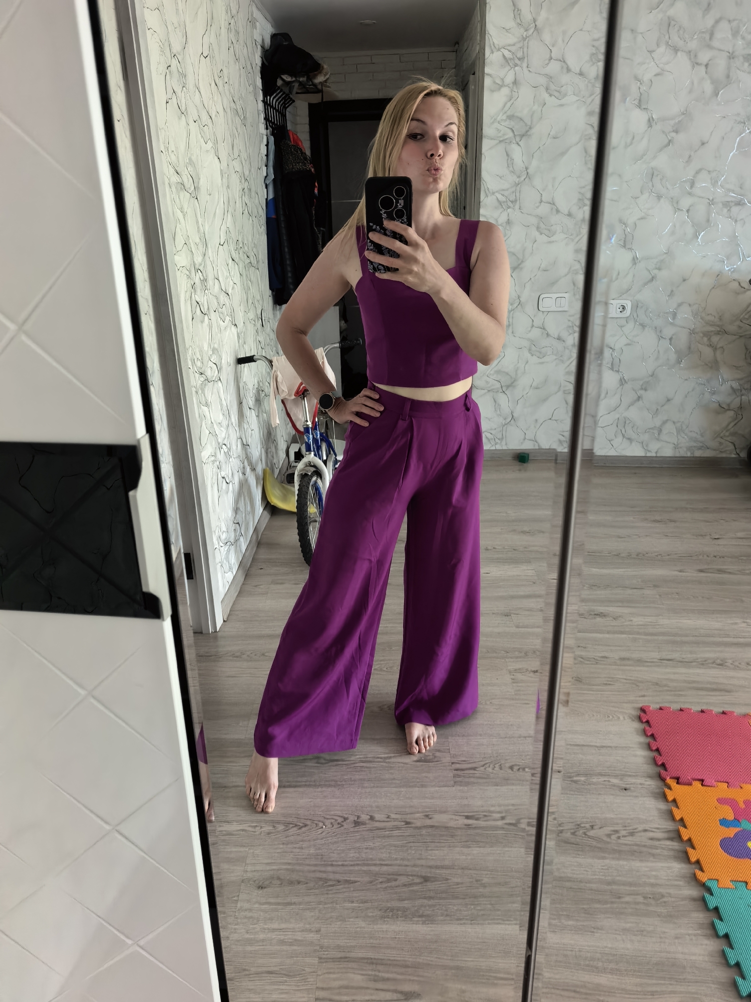 A woman with blonde hair is standing in front of a mirror, taking a selfie. She is wearing a matching purple tank top and high-waisted pants. Her right hand is on her hip and she has a pursed lip expression. The woman is in an interior space. Behind her, against the wall is a child's bicycle, and also a hanging rack with clothes. Further back, the wall has a marble-like pattern. The floor is wooden. A play mat is visible on the floor to the right.