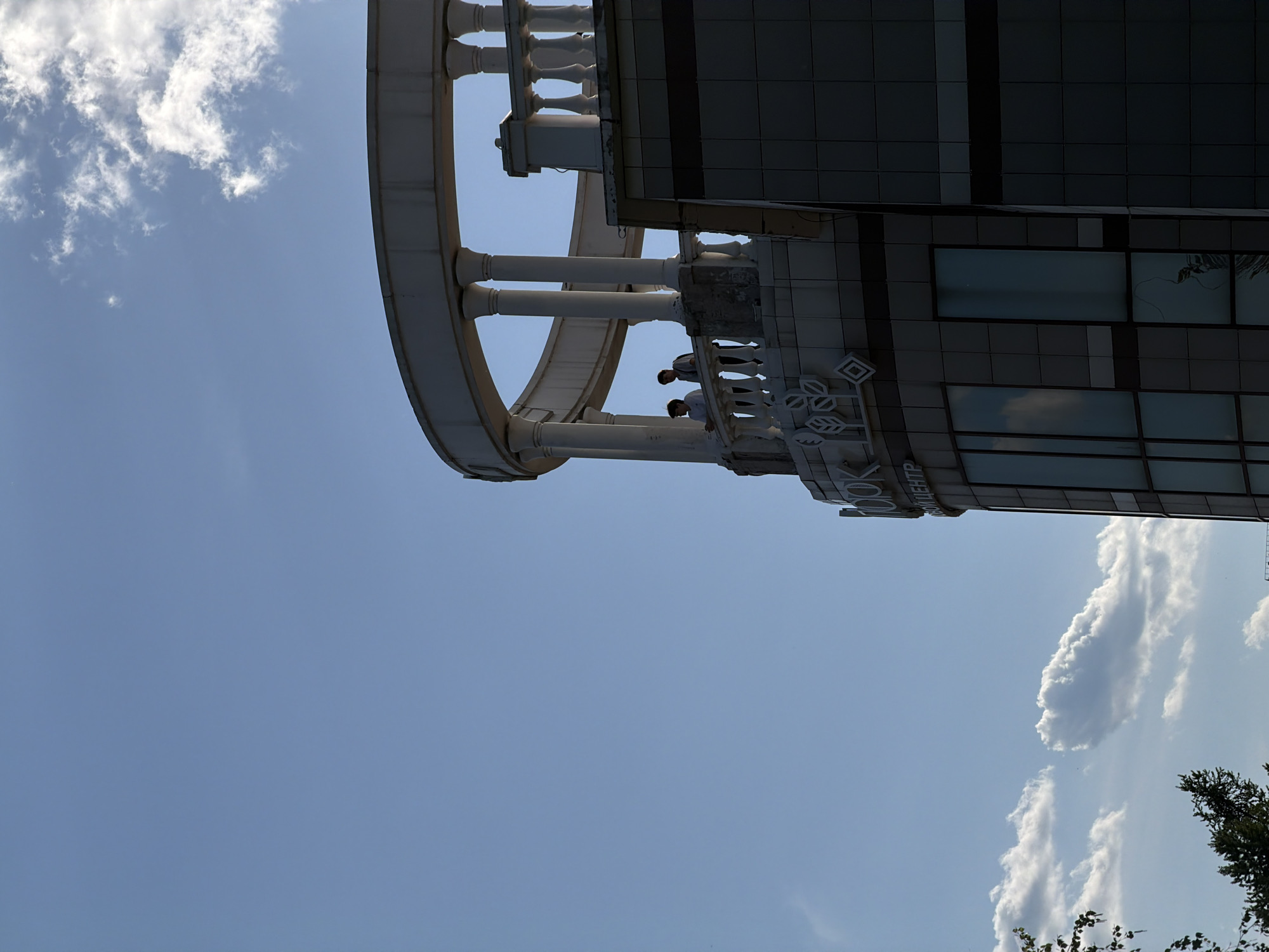 The image depicts the Krasnoyarsk Regional Philharmonic building in Krasnoyarsk, Russia. A portion of the building is visible against a blue sky with scattered clouds. The architectural details of the building include a curved balcony with columns and a balustrade. Two people are standing on the balcony. The building's facade has a sign with Russian text, and the lower part of the image shows parts of a tree.