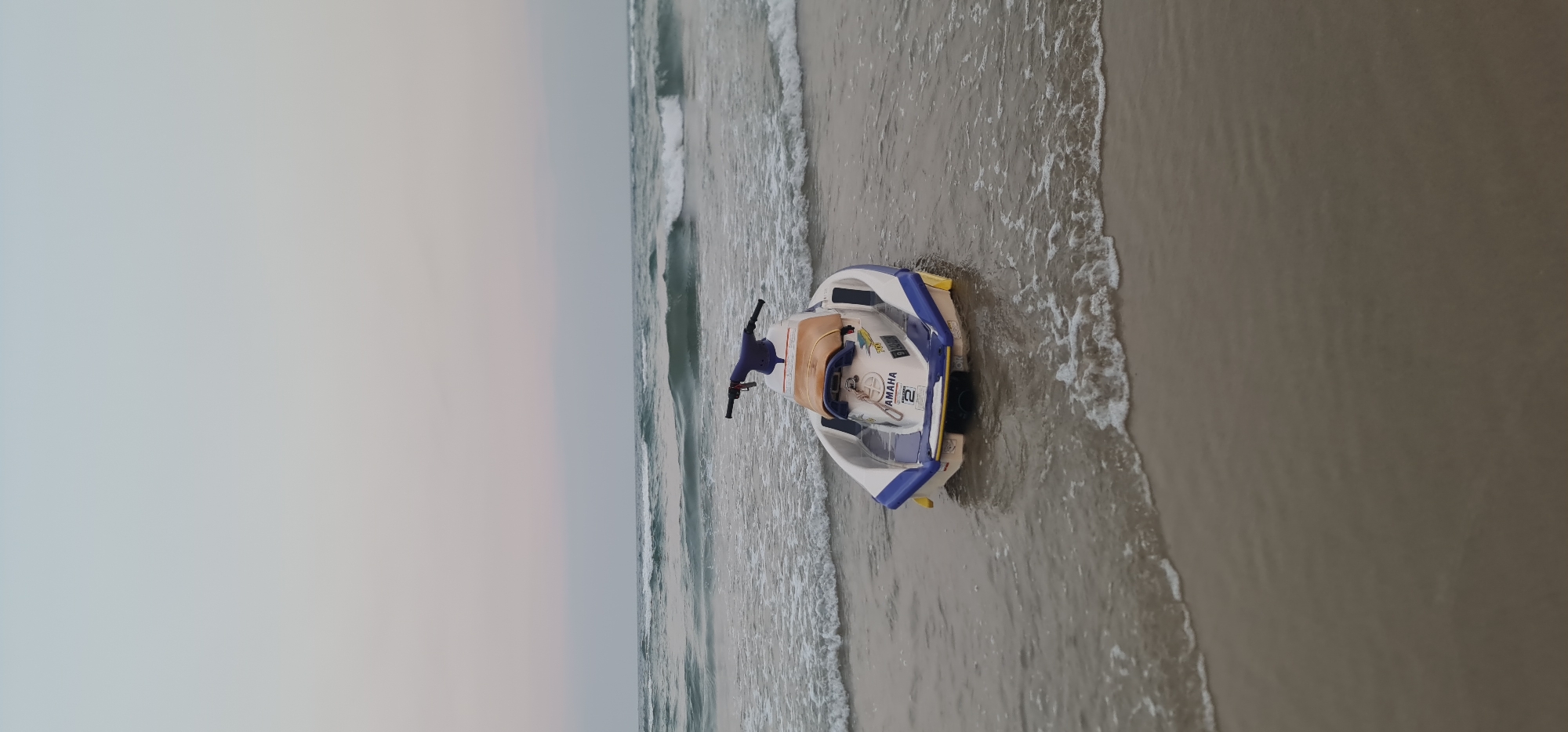 A white and blue Yamaha WaveRunner personal watercraft is beached on the shore. The sky is overcast and gray. Waves are visible in the ocean. The sand is beige and wet. The location is a beach in Đà Nẵng, Vietnam. No people are visible.