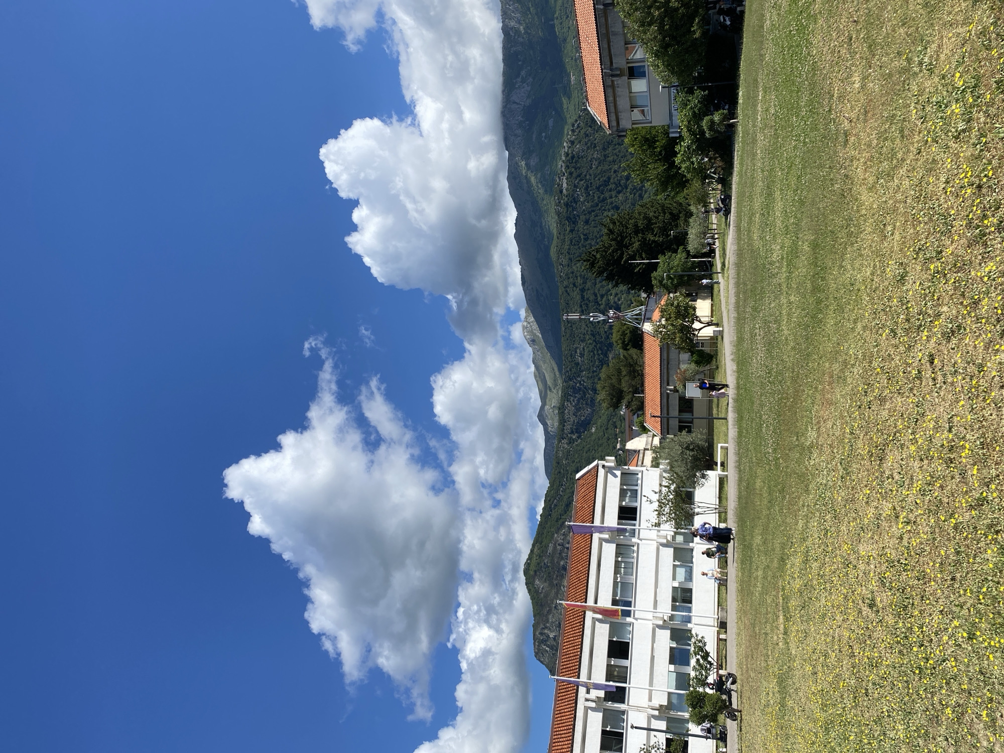 The scene is an outdoor landscape in what appears to be Budva, Montenegro, featuring a clear blue sky with puffy white clouds, a mountain range, buildings, and a grassy field.
On the left side of the image, white buildings with orange tile roofs are visible. Flags are mounted on the facade of the buildings.
The mountain range rises in the background, covered with green vegetation. A radio or communication tower is discernible near the base of the mountain.
On the right side of the image, a grassy field is filled with small yellow flowers. People can be seen walking near the buildings along a paved path. The photograph is taken during the daytime, with bright sunlight illuminating the scene.