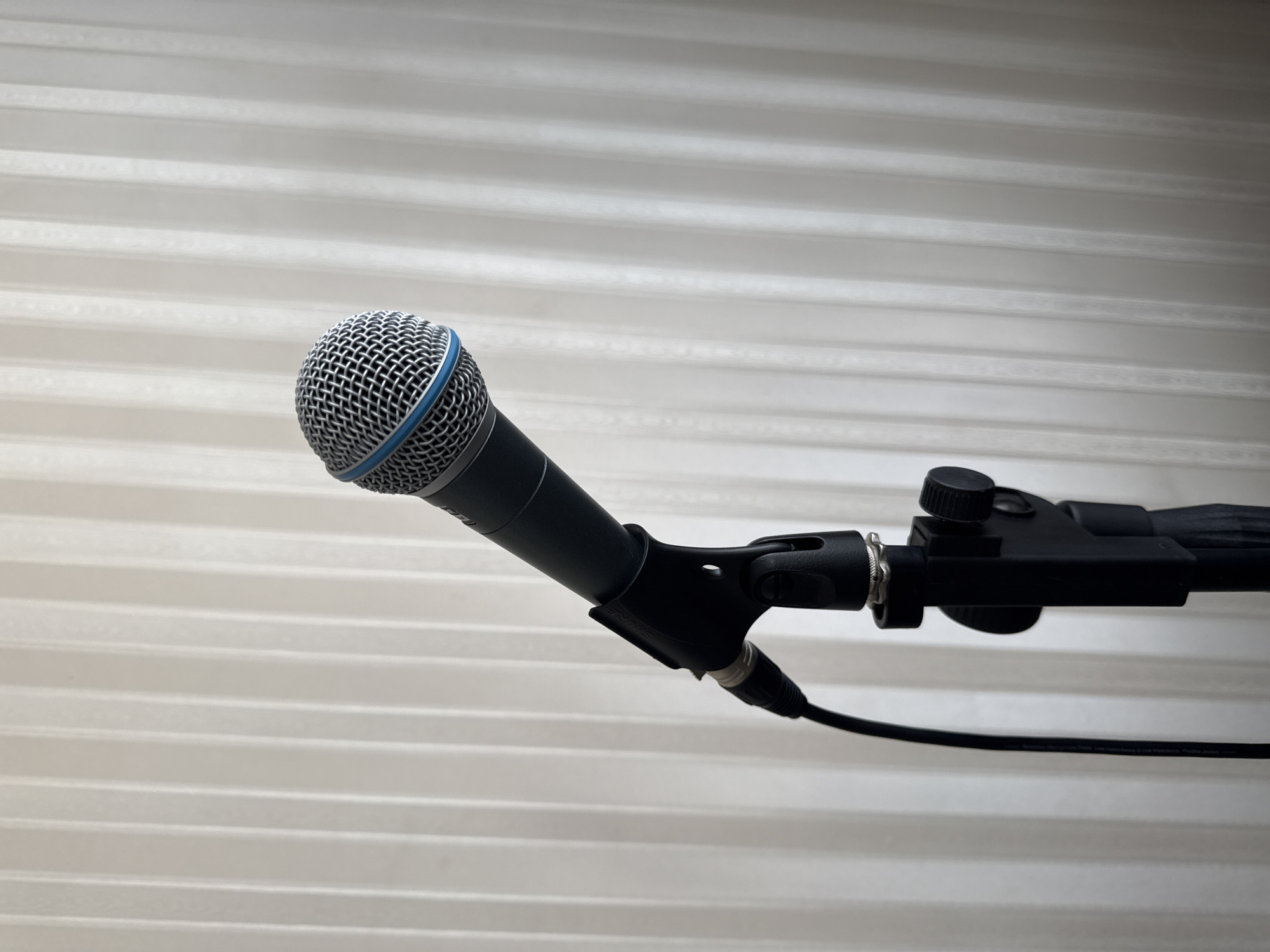 A close-up, slightly angled shot shows a black microphone on a microphone stand. The microphone is dark grey with a silver grille and a blue accent ring. A black cable connects to the bottom of the microphone. The stand is black and appears to be made of metal. The background is a pale, off-white surface with vertical pleats or folds, resembling blinds or curtains. The lighting is soft and diffused. There are no people, animals, or other objects visible in the frame. The image does not provide any information about its location in Plovdiv, Bulgaria.