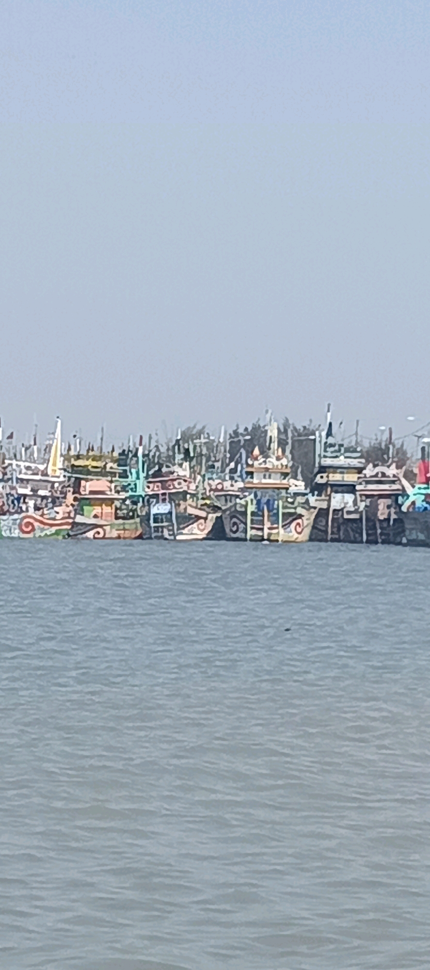 The image shows a fleet of vibrantly painted fishing boats docked in the waters of Blimbing, Indonesia. The boats are lined up closely together, forming a colorful spectacle against the hazy, pale blue sky. Many of the boats feature intricate, swirling patterns in various colors, including red, orange, yellow, green, and blue, with some also adorned with what appear to be decorative structures resembling small pagodas or shrines on their decks. The water is a calm, muted gray-blue, with gentle ripples suggesting a tranquil environment. The background is dominated by the sky, with the distant shoreline or landmass barely visible as a dark, fuzzy line obscured by haze and the density of the boats. There are no people visible in the frame, and the focus is entirely on the static display of the decorated vessels.