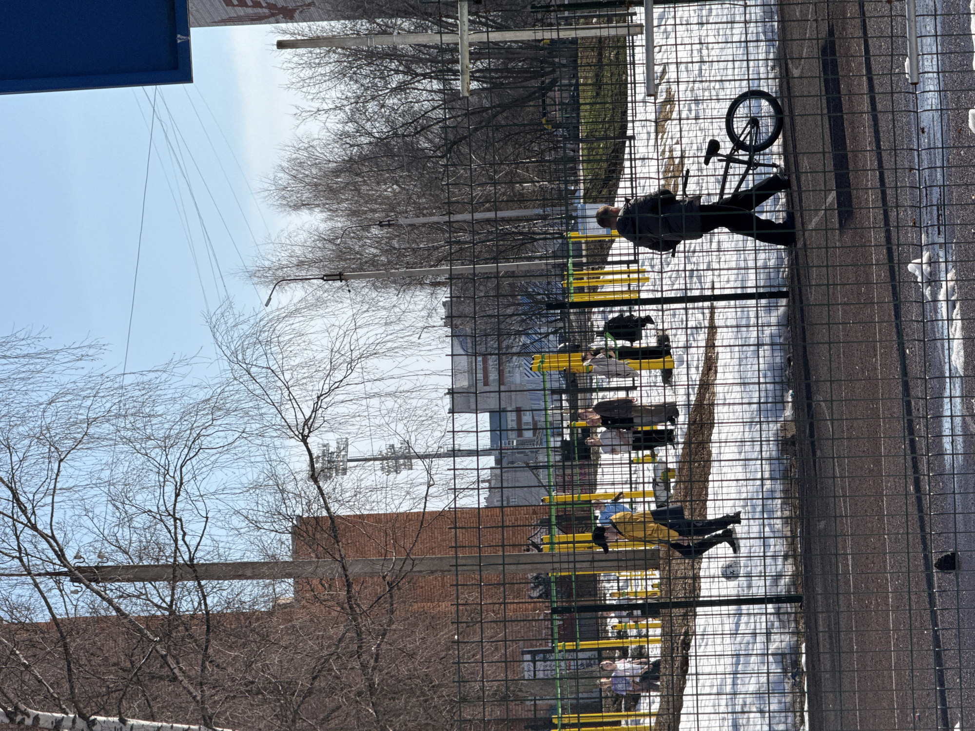 The image depicts a scene in Votkinsk, Russia, as evidenced by the typical Soviet-era architecture of the building in the background. The scene shows a group of people gathered near a metal fence, possibly a playground or recreational area. Some individuals are standing and appear to be conversing, while others are seated on yellow benches. A bicycle is leaning against the fence near the edge of a paved area. Patches of snow are visible on the ground, suggesting a late winter or early spring setting. The environment includes bare trees, indicative of the season, and glimpses of other buildings beyond the immediate area. The scene appears to capture a typical urban recreational space in Votkinsk.
