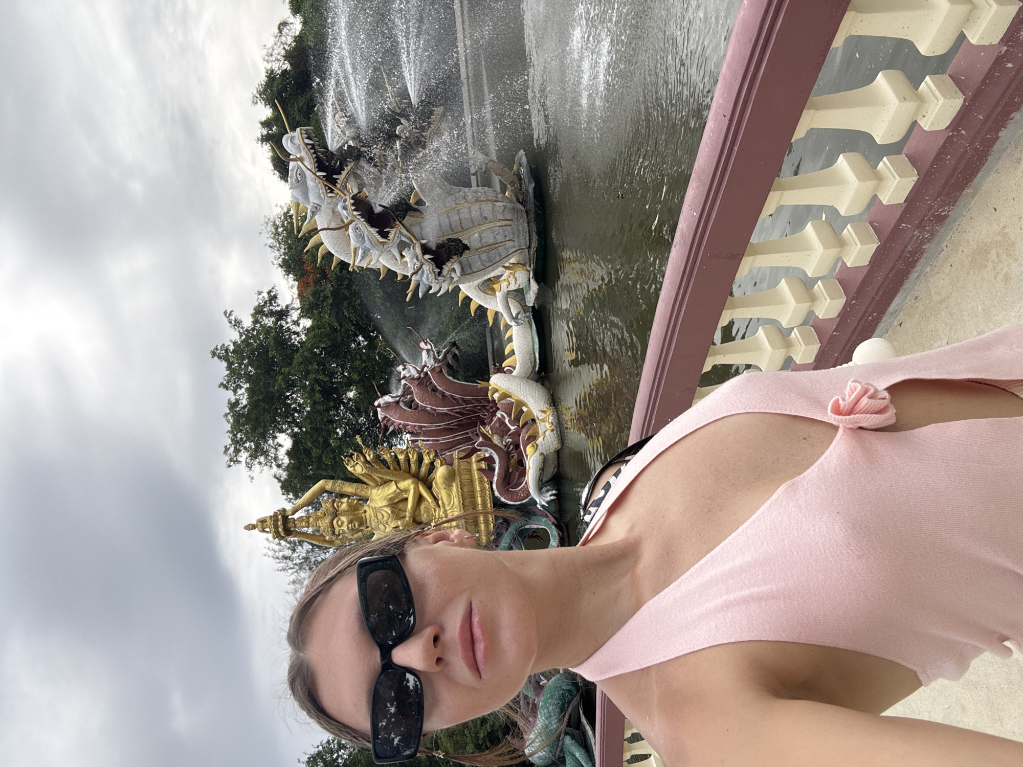 In Bang Pu Subdistrict Municipality, Thailand, a young Caucasian woman with blonde hair, wearing black rectangular sunglasses and a pink halter top, stands in the foreground and takes a selfie. Behind her, a pond with a fountain featuring multiple dragon heads spewing water, and a golden statue of a multi-armed deity, are visible. The woman is positioned on a walkway with a decorative railing.