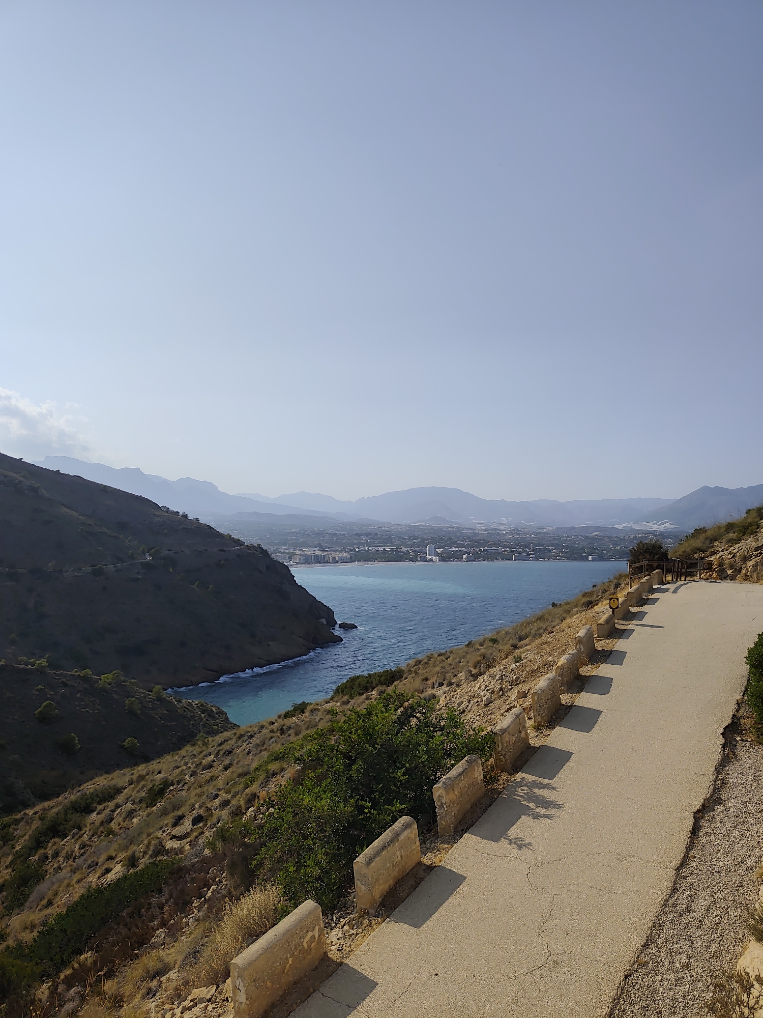 The image shows a panoramic view of a coastal area, likely in or near l'Alfàs del Pi, Spain. A paved road curves along the right side of the frame, bordered by a low stone wall. The road is surrounded by dry, scrubby vegetation and rocks. The road leads towards a bay with vibrant blue water. In the distance, across the bay, is a coastal town with buildings and mountains rising behind it. The sky is a clear, pale blue. The overall impression is of a scenic, elevated viewpoint overlooking the Mediterranean Sea.