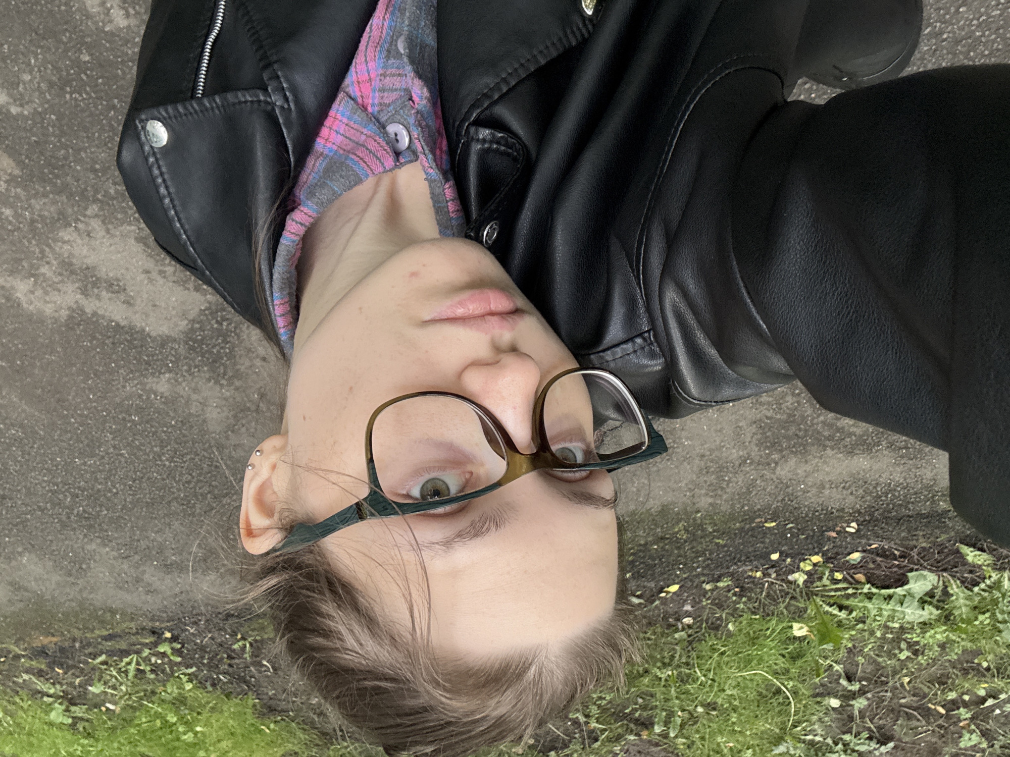 The image shows a close-up, sideways view of a person wearing glasses, a pink and grey plaid shirt, and a black leather jacket. The person's face is partially visible, with one eye looking towards the camera. The background consists of a patch of green grass and a grey, textured surface that appears to be asphalt or a similar urban ground material. The lighting suggests an overcast or shaded environment. The image is oriented such that the top of the person's head is on the left side of the frame, and their feet are on the right. There are no discernible Saint Petersburg landmarks or specific contextual clues to place the location within the city.