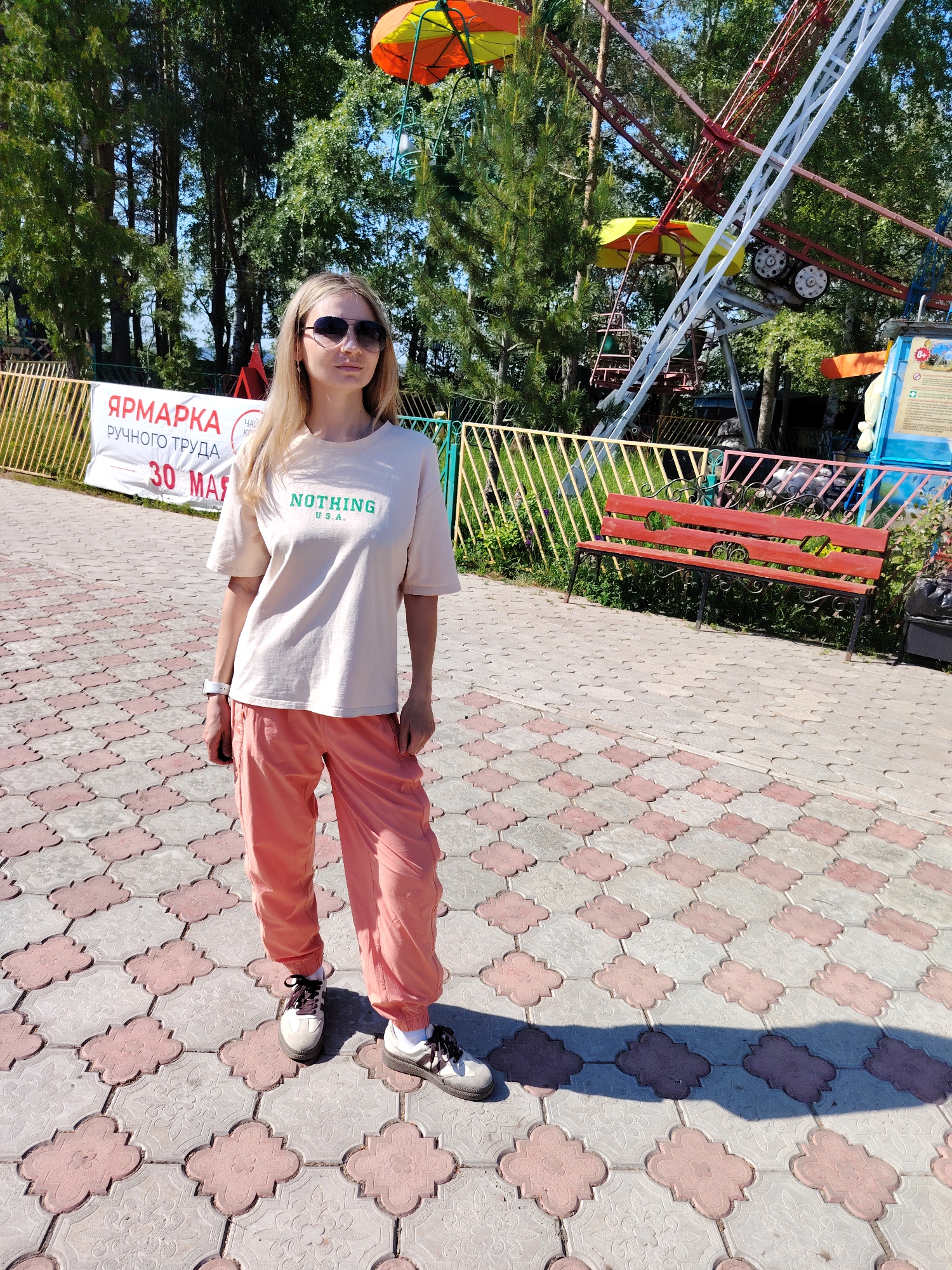 A woman stands on a paved walkway. She has long blonde hair and wears sunglasses, a beige t-shirt with "NOTHING USA" printed in green, and salmon-colored pants. She is wearing white socks and sneakers. In the background is a fence and some trees, along with the partially visible structure of an amusement park ride. Behind the woman is a sign that reads "YARMARKA RUCHNOVO TRUDA 30 MAI" (translated: HANDICRAFT FAIR MAY 30th). A red bench sits next to the walkway. The location is the city of Chaikovsky, Russia.