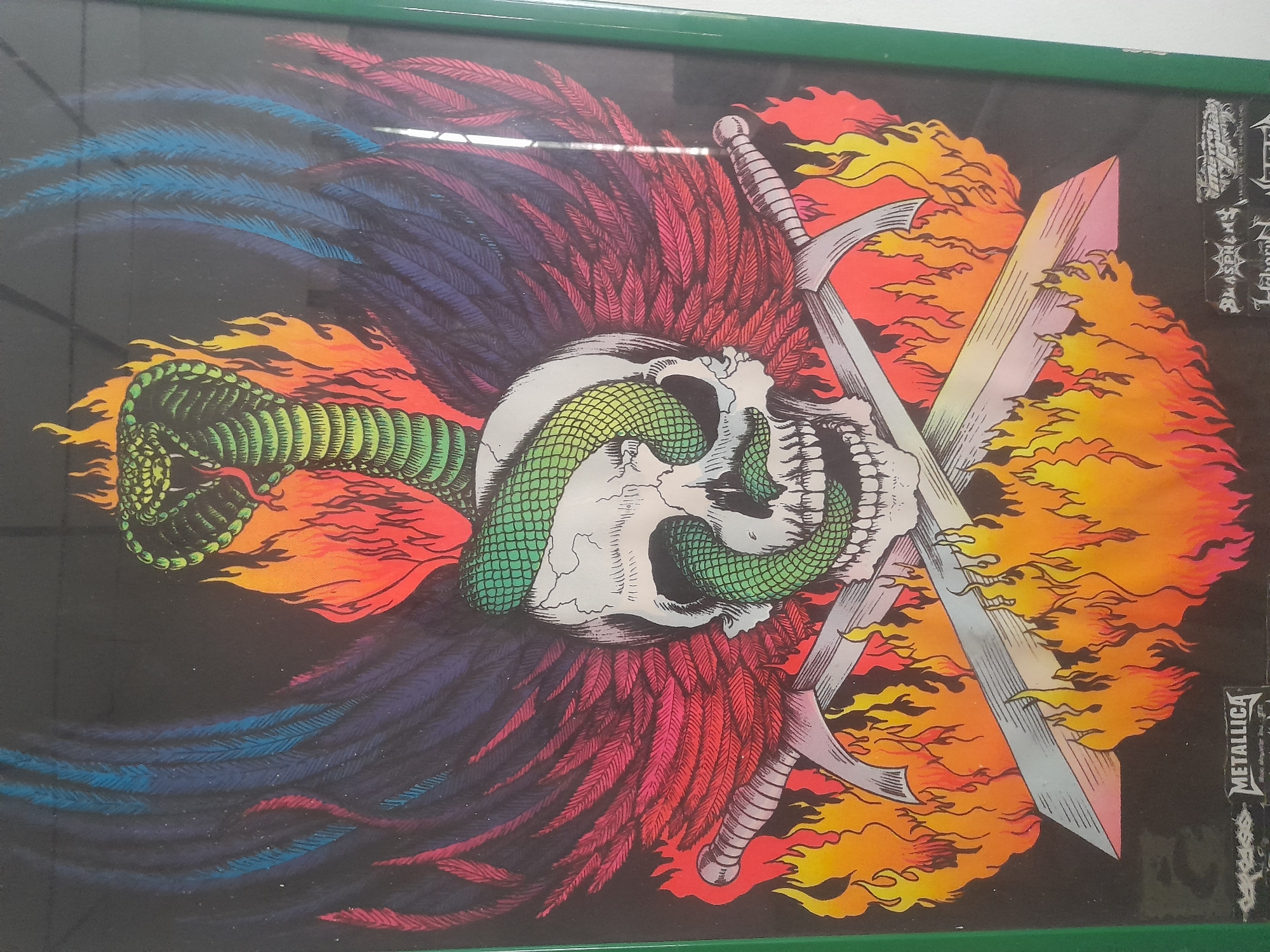 Framed artwork depicting a skull with a snake wrapped around it, feathers, and crossed swords with flames. The artwork also has text on the right side that reads "METALLICA" and "BLASTERED". The frame is green. The location is Tinaquillo, Venezuela.
