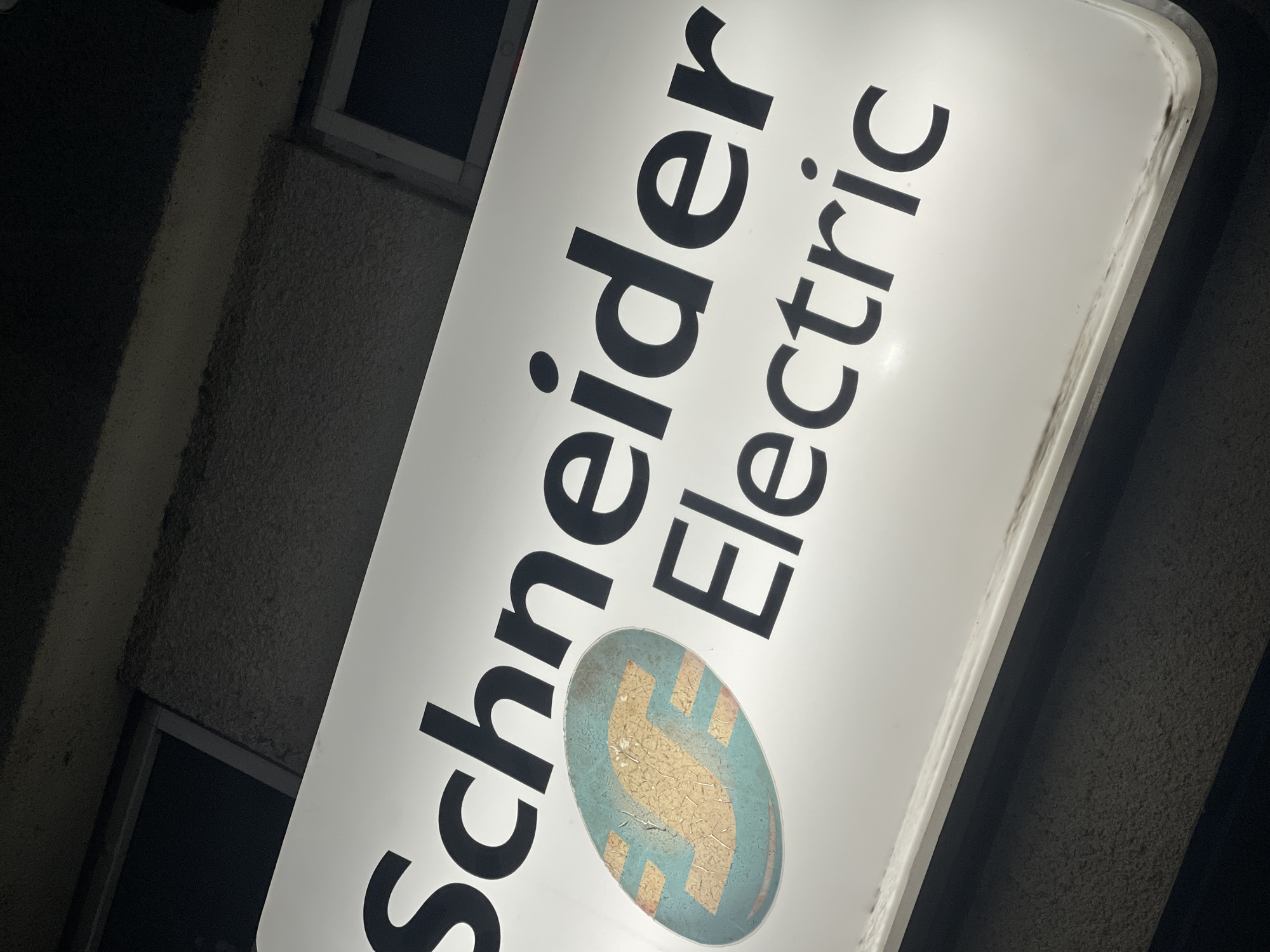 The image shows a Schneider Electric sign, illuminated and affixed to a building exterior. The text "Schneider Electric" is displayed prominently in bold, black lettering on a white background. Below the text is an oval logo with a stylized "S" design, appearing in teal and gold. The sign is mounted at an angle. The building's exterior appears to be textured and of a dark, possibly gray color. A small window is partially visible behind the sign. No people or interactions are observed in the image. The image was taken in Burgas, Bulgaria.