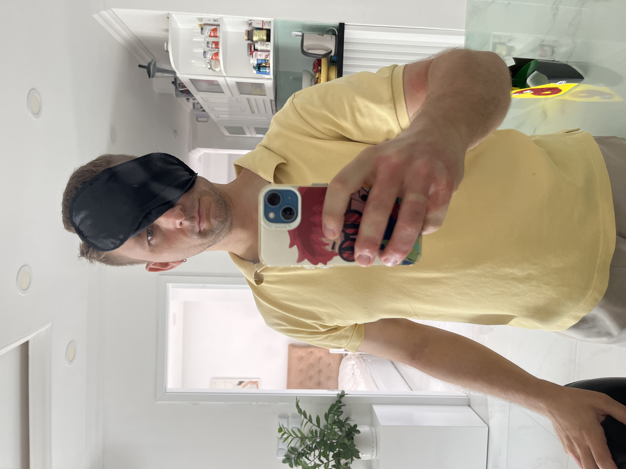 A man is taking a selfie in a mirror. He is wearing a black sleep mask over his eyes and a yellow collared shirt. He is holding a phone with a white and patterned case. The reflection in the mirror shows a kitchen with white cabinets and a white countertop. A potted plant is visible on the counter near the bottom of the frame. A doorway behind the man leads to another room. The location is implied to be in Bac Nha Trang Ward, Vietnam.