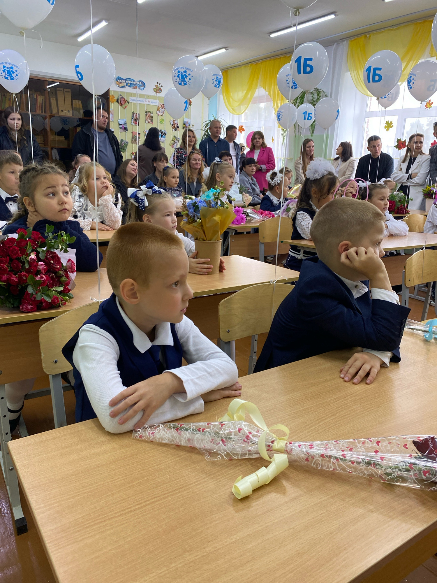 The image depicts a classroom setting, likely for a school event, as indicated by the presence of balloons with the numbers "15" and "6" and "1", and floral arrangements. The room appears to be decorated with festive elements. In the foreground, two young boys are seated at wooden desks, facing towards the right of the frame. The boy on the left is wearing a white collared shirt and a dark blue vest, with his left hand resting on his chin. The boy on the right is similarly dressed in a white shirt and dark blue jacket, with his right arm resting on the desk.
Behind the boys, several children, predominantly girls, are also seated at desks. They are dressed in school uniforms, many with white blouses and dark skirts or vests, and some have elaborate hair decorations. Adults are visible in the background, standing or seated, observing the children. The environment is a classroom with desks and chairs arranged in rows. The walls are decorated with posters and what appear to be photographs. Sunlight is entering the room, illuminating the scene. The presence of balloons and flowers suggests a celebration or the start of the school year. The text on the balloons appears to be in Russian, with one balloon showing "ЗДРАВСТВУЙ ШКОЛА" (Hello School) and other balloons with numbers. The overall atmosphere is one of a formal gathering or event. The location is identified as Noviy, Russia, based on the prompt.