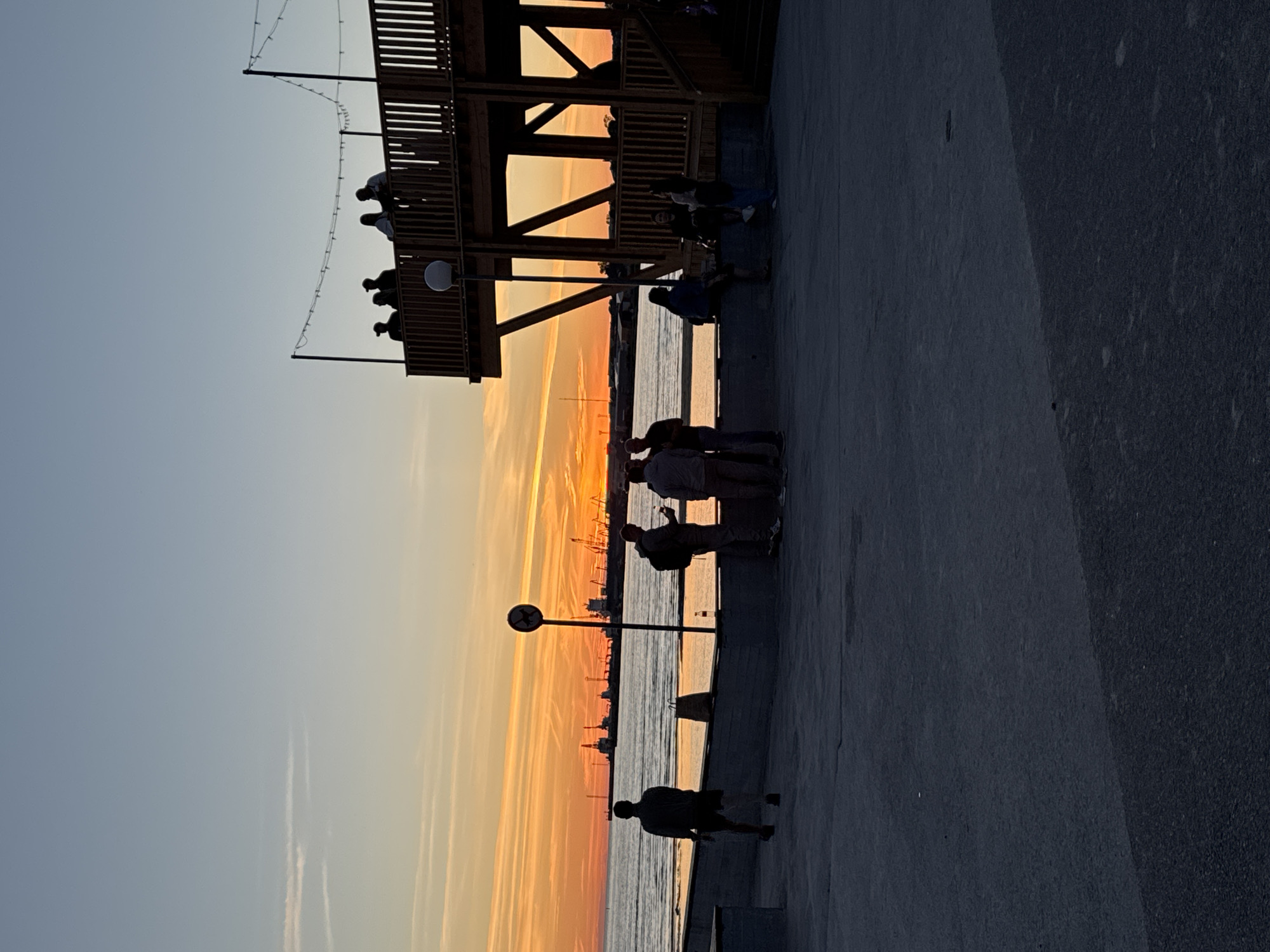 The image depicts a twilight scene at the Tallinn coastline, likely near the Linnahall or a similar waterfront area in Tallinn, Estonia. The sky displays a gradient of colors, transitioning from a pale blue at the top to vibrant orange and yellow hues near the horizon, indicative of a sunset or sunrise. Silhouetted against the colorful sky are several individuals.
In the foreground, a person is walking away from the camera, facing towards the water. To the right, a group of people are gathered, some standing and conversing, while others are seated. Behind them, a multi-level wooden structure, possibly a viewing platform or cafe, rises into the frame, adorned with string lights. In the middle ground, the sea stretches out, reflecting the sky's colors. Distant industrial elements, such as cranes and ships, are visible on the horizon, suggesting a port or industrial zone. A circular sign with a silhouetted dog and a diagonal line is prominently displayed on a pole near the water's edge. The overall atmosphere is serene, with people enjoying the coastal scenery at dusk.