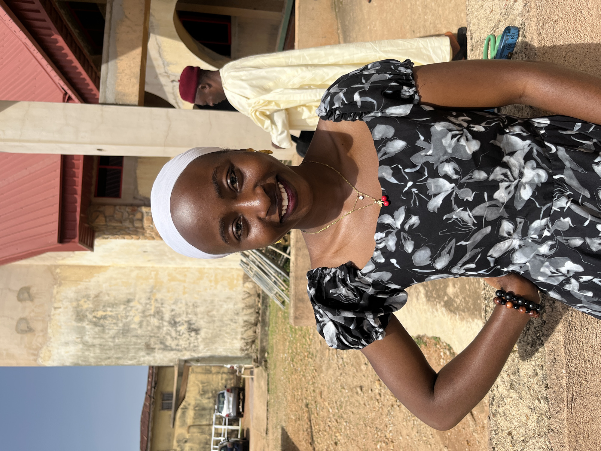 In the city of Bukuru, Nigeria, a young Black woman wearing a white headscarf and a black and gray floral dress stands facing the camera. Her left hand is on her hip, and she is smiling broadly, revealing white teeth. A delicate gold necklace with a red cherry pendant hangs around her neck. To the right and behind her, an older Black man in a light yellow robe and a red cap is visible from the waist up, walking away from the camera. The background features a building with a red roof and beige walls, suggesting an outdoor, possibly religious or community setting. The ground is covered with dirt and scattered debris, and in the distance, cars are parked. A sewing tape measure is on the ground to the right of the woman.