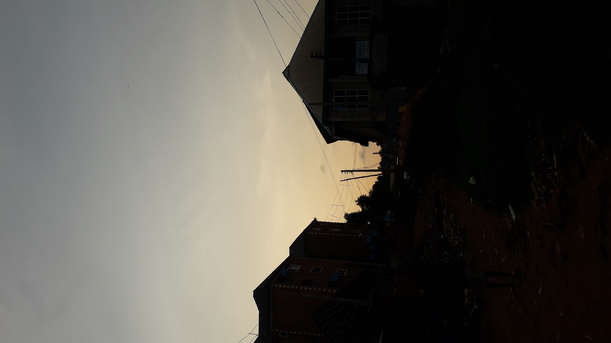The image shows a street scene in Nkpor, Nigeria at dusk. Two buildings are silhouetted against the fading light, one on either side of the street. Electrical wires are visible overhead. The road appears unpaved and dark. Details in the foreground are obscured by shadow. No people are discernible.