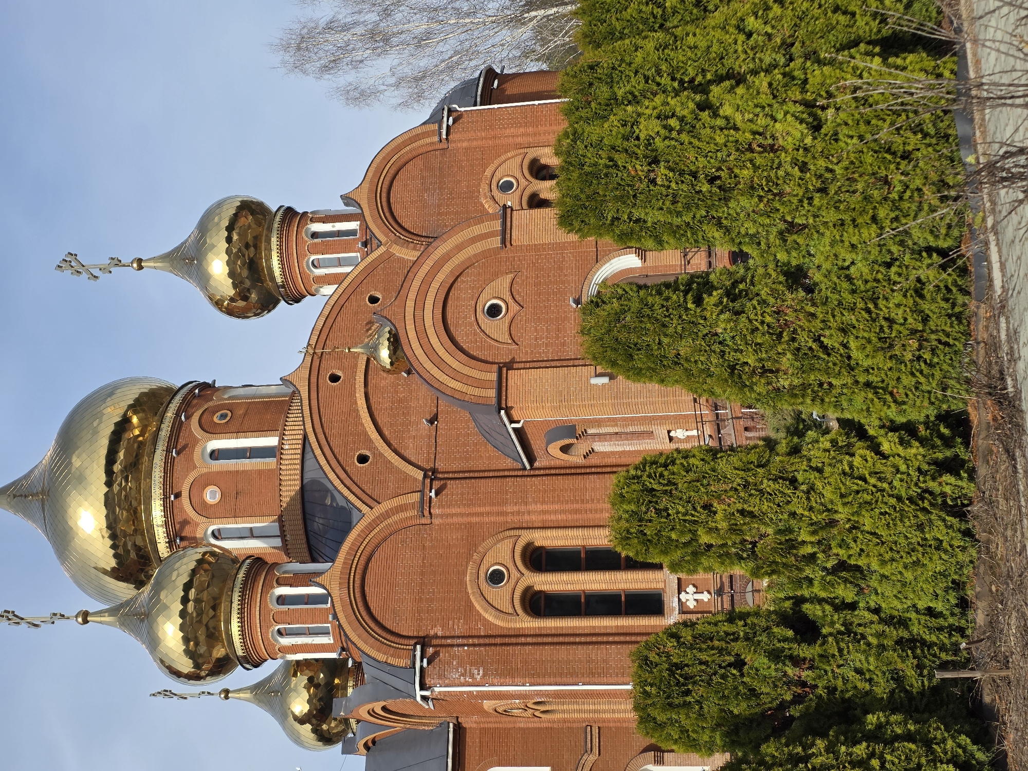 The image shows the Cathedral of St. George the Victorious in Chaikovsky, Russia. The red brick building has multiple golden onion domes topped with Orthodox crosses. Green trees partially obscure the facade. The sky is light blue.