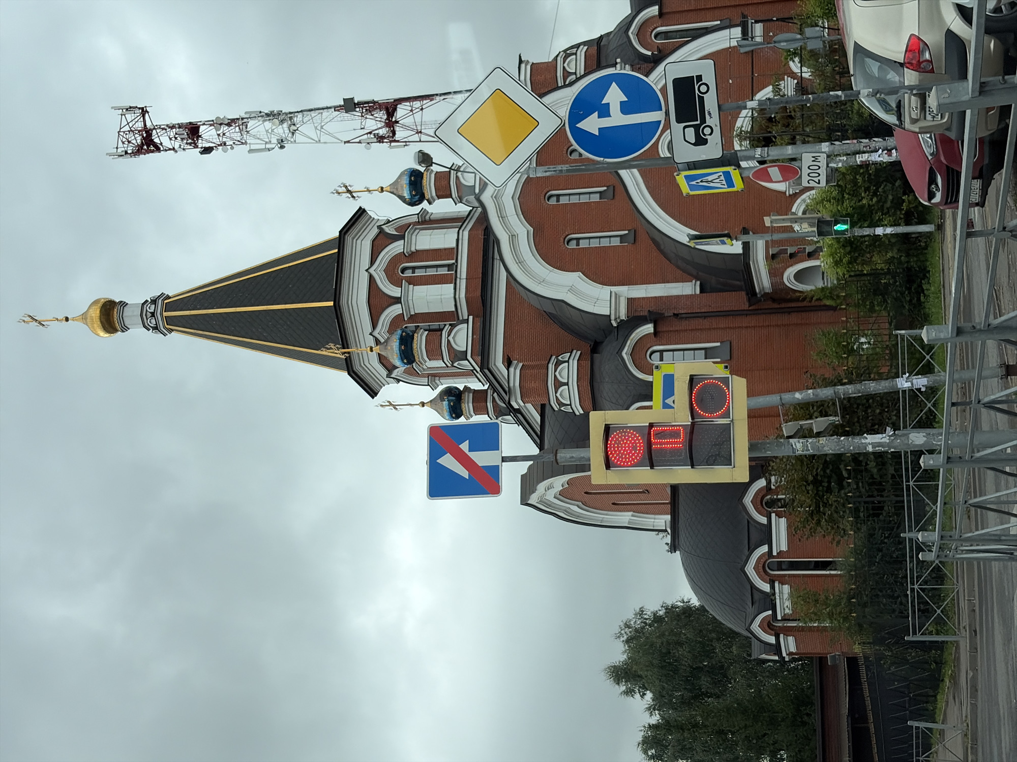 The image depicts a street scene in Balashikha, Russia, featuring a red brick church with a golden dome and blue accents. The church has arched windows and is partially obscured by trees and a metal fence. Several traffic signs are visible, including a "no entry" sign, a directional sign indicating straight ahead only, a "priority road" sign, a pedestrian crossing sign, a truck route sign, and a traffic light showing red and amber lights, with a green light visible further down. A cell tower stands tall in the background. Two cars, a red sedan and a beige hatchback, are parked on the street. The sky is overcast, indicating a cloudy day.