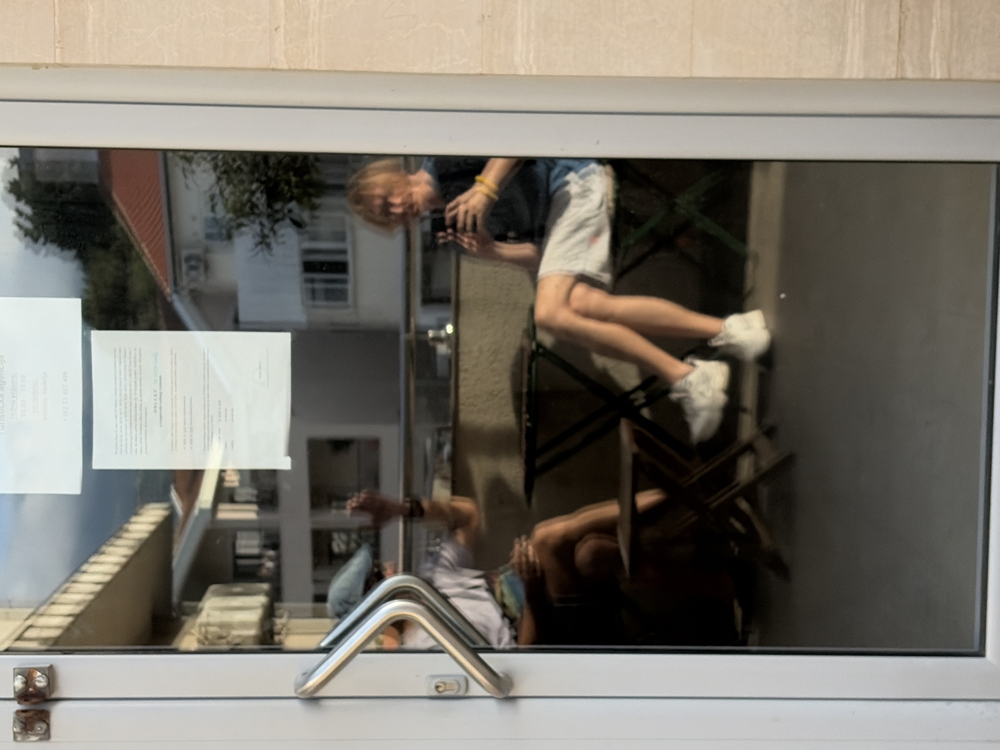 The image captures a reflective window, likely from a shop or building entrance, framed by a white window structure and a beige-tiled wall above. The photograph itself is rotated 90 degrees clockwise, presenting the scene sideways.
The dominant subject is the reflection of a person, appearing to be a young individual with light hair, wearing a dark t-shirt, light shorts, and white sneakers. If the image were correctly oriented, this person would be seated on a dark, possibly foldable, chair, holding a black smartphone with both hands, seemingly taking a picture or looking at the screen. Their head is visible on the left side of the reflection (top if un-rotated), and their legs, bent at the knees, extend towards the right (bottom if un-rotated).
Through and around the main reflection, elements of the outdoor environment are visible: green trees, parts of a building with a red roof, windows, and an air conditioning unit. Another fainter reflection of a person, possibly seated and wearing a hat, is visible closer to the bottom of the window (left if un-rotated).
Two pieces of paper are taped to the inside of the window, visible through the reflections. The left paper clearly shows the word "agencija" and partially "turistička", suggesting it's an advertisement or information for a "tourist agency." The text on the right paper is less discernible.
The foreground shows the window frame's metallic handle, shaped like a 'V' when closed, and two small, square, metallic hinges on the bottom-left corner of the frame. The scene appears to be set during daytime under bright conditions, indicated by the quality of light and reflections. Given the location context of Budva, Montenegro, the "tourist agency" text is fitting.